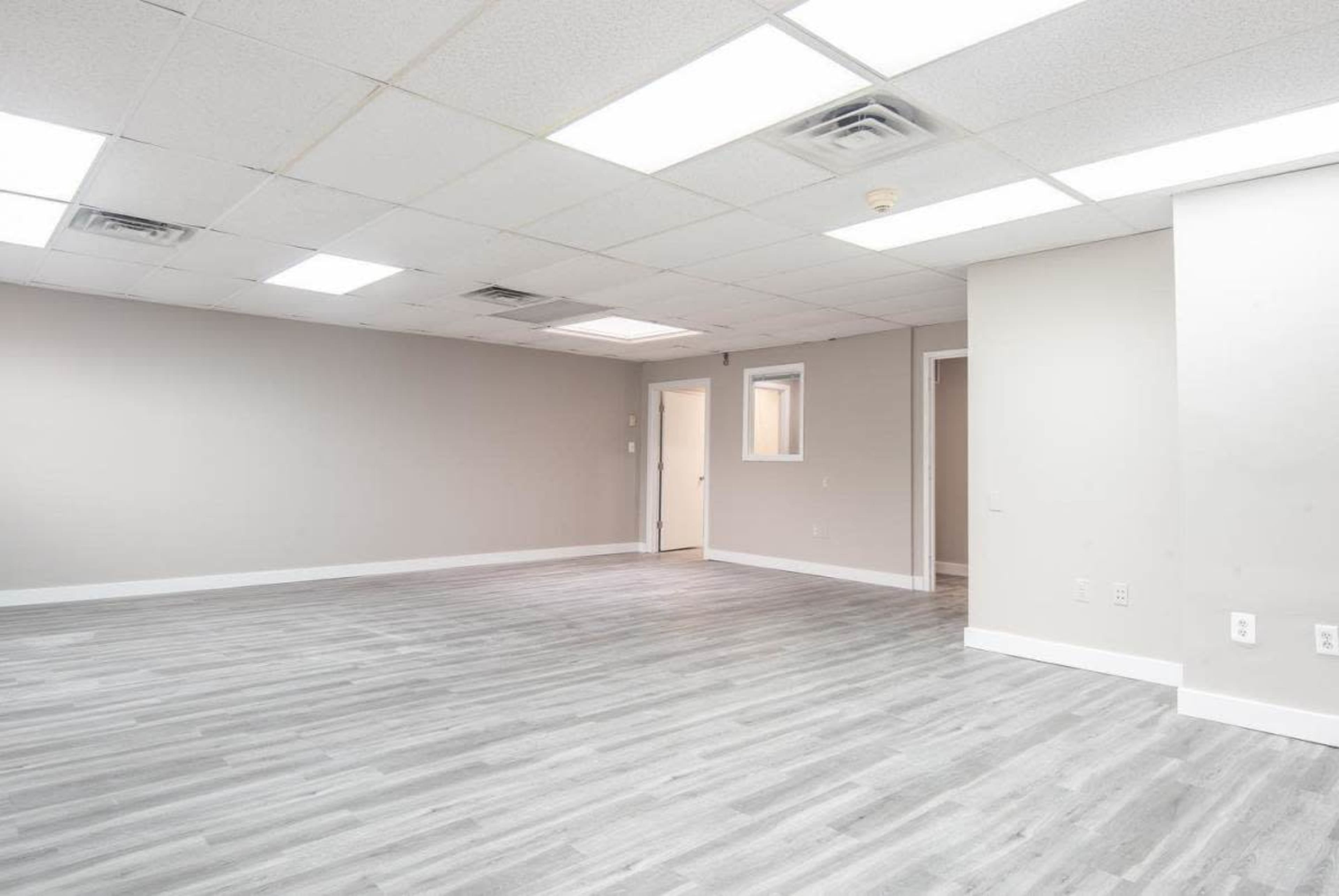 A vacant room with light gray flooring and neutral-colored walls, featuring a ceiling with fluorescent lights and a doorway leading to another space.