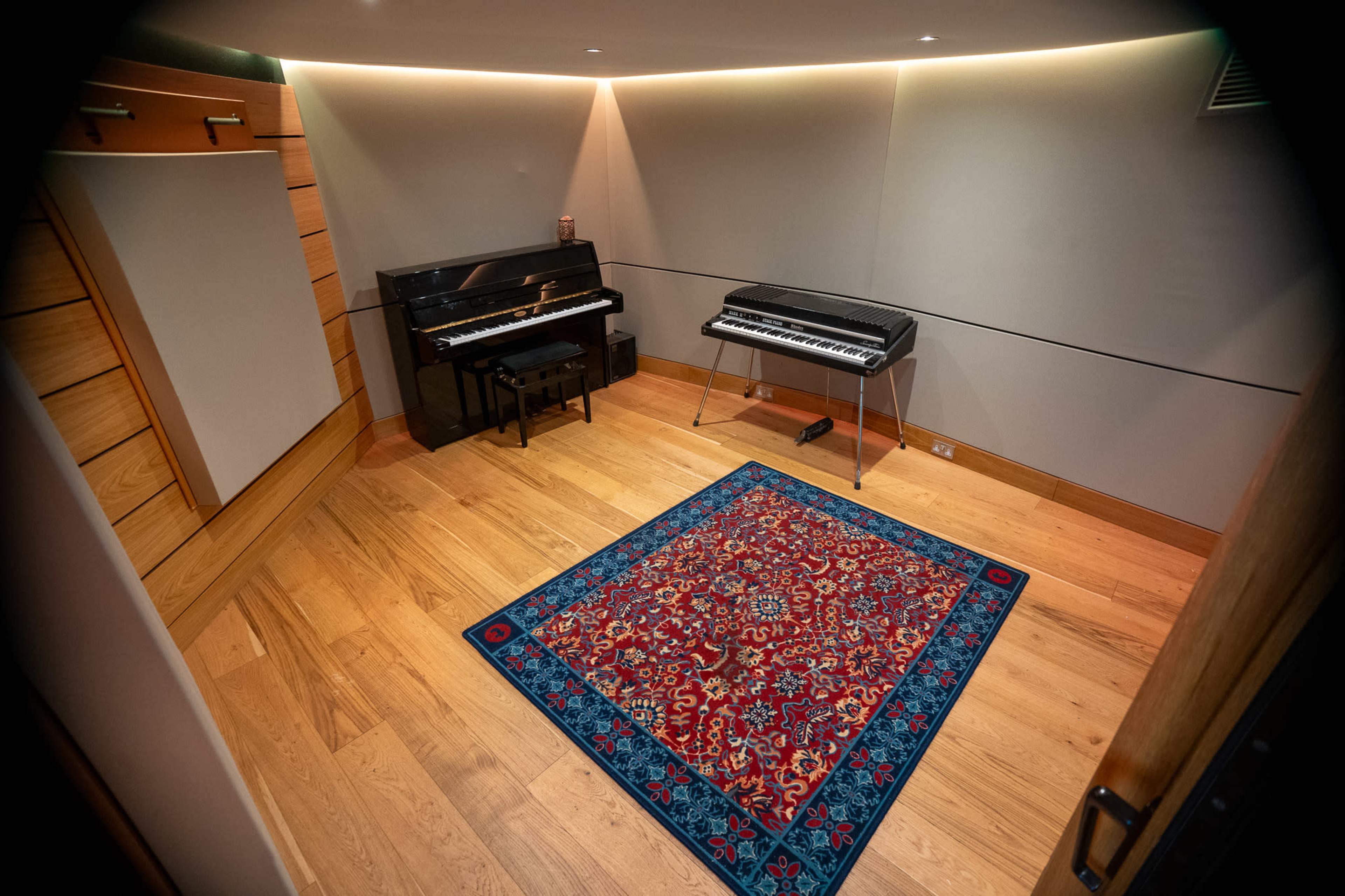 The image shows a well-furnished room with wooden flooring, featuring a black piano and a keyboard on stands, accompanied by a decorative rug in the center.
