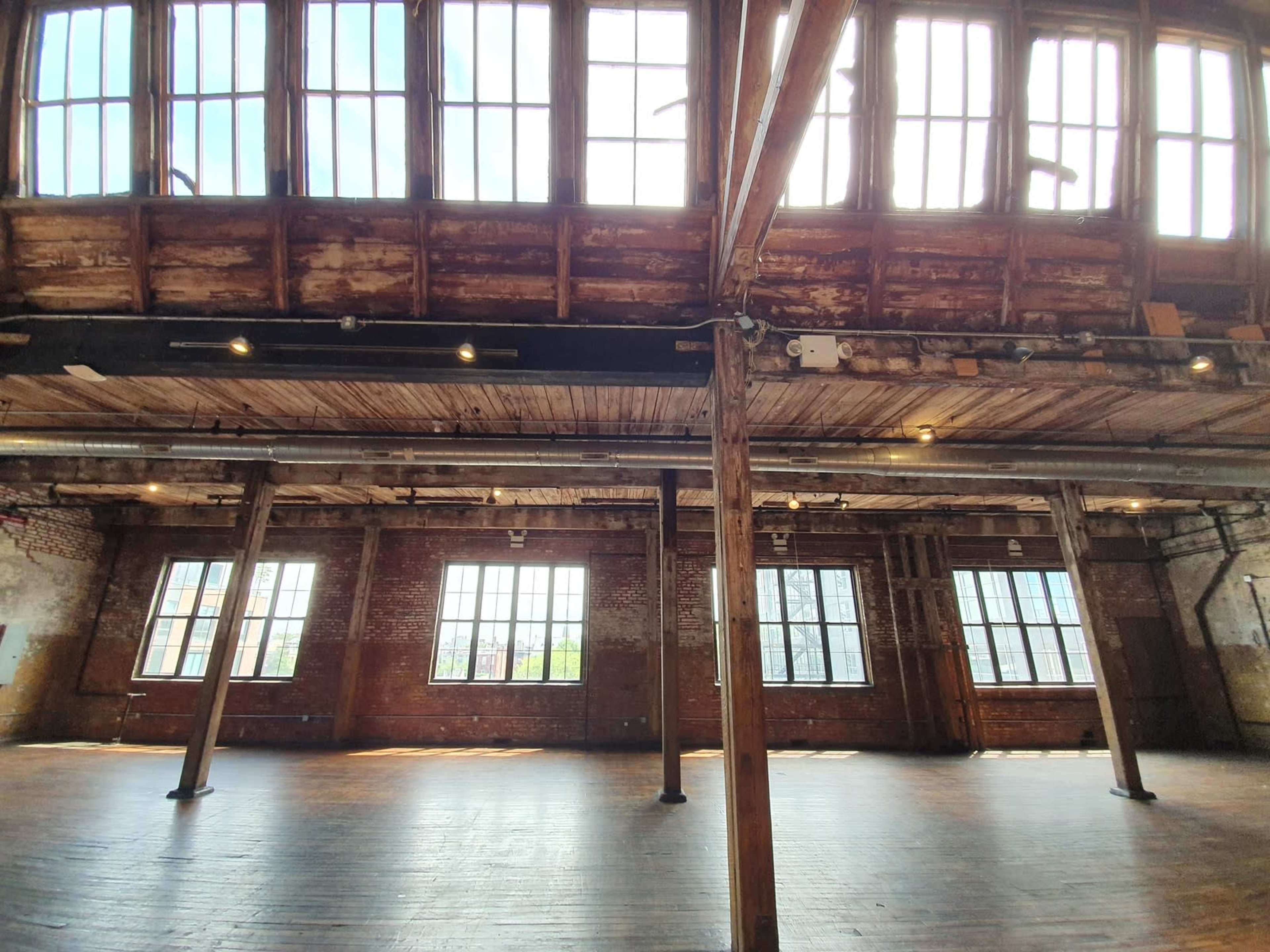 An empty industrial space with large windows and exposed wooden beams, featuring wooden floors and a high ceiling.