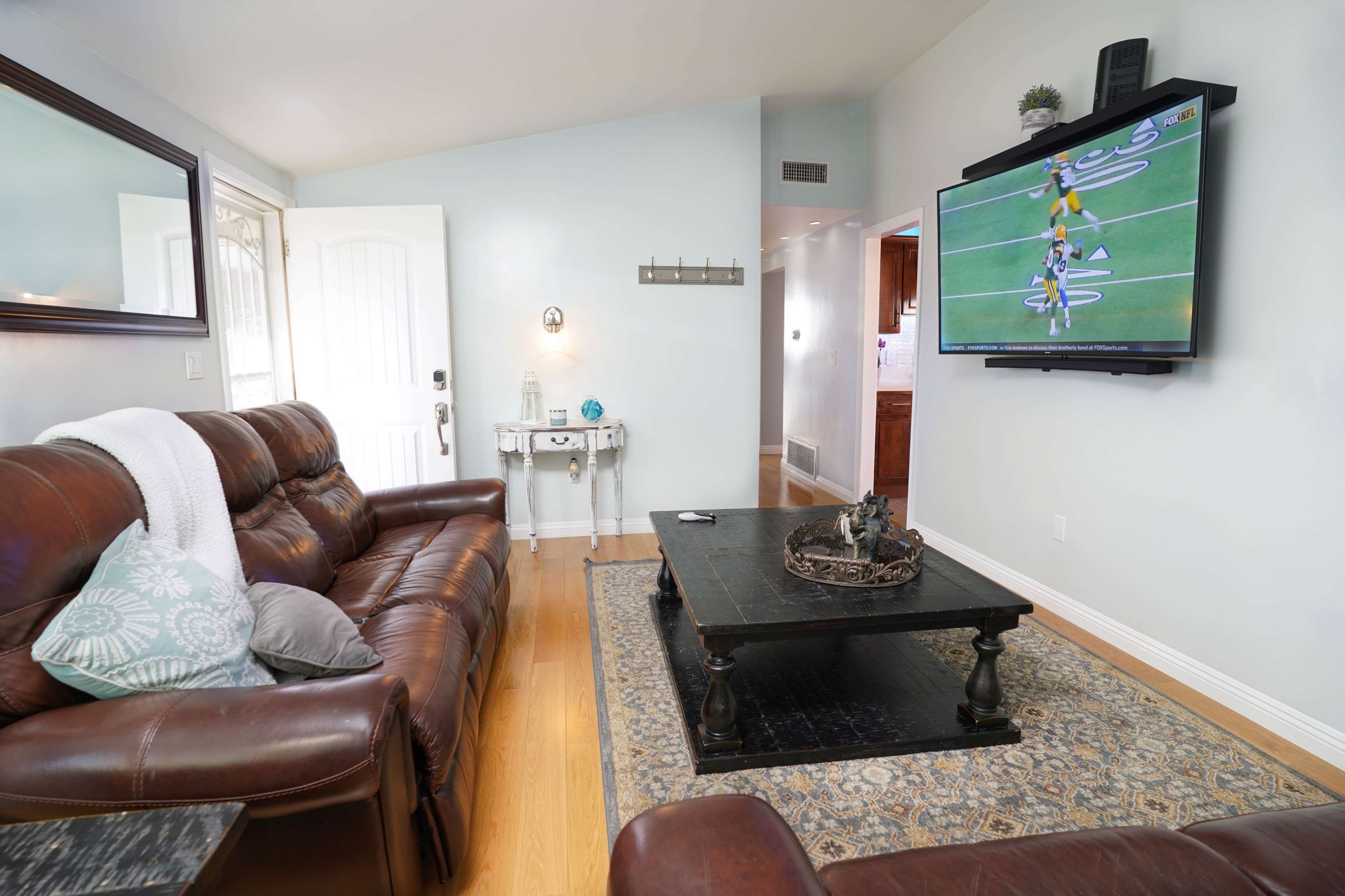 The living room features a brown leather sofa, a dark wooden coffee table, and a flat-screen television mounted on the wall, with a door visible in the background.