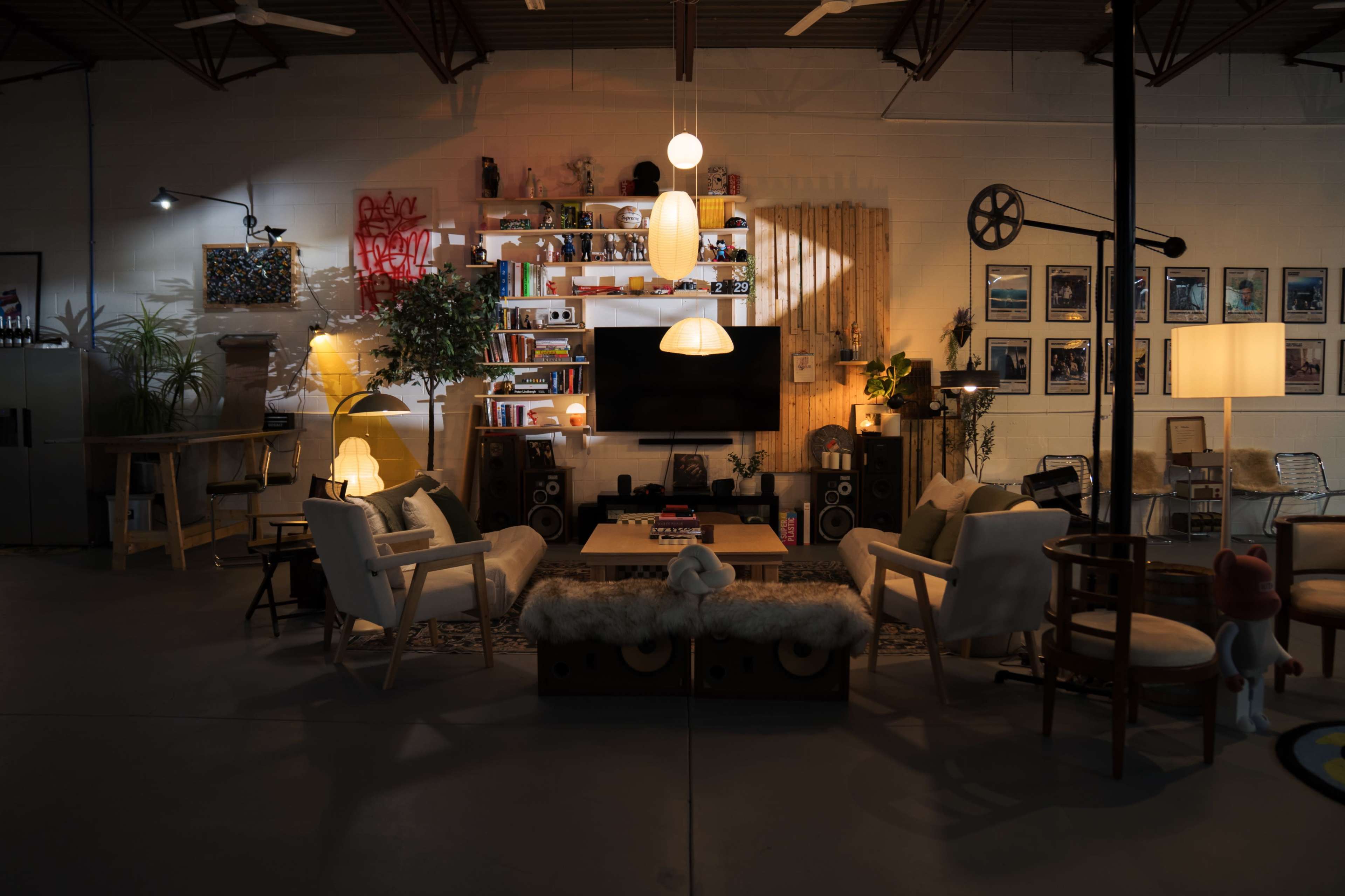 The image shows a modern living space with a mix of furniture, featuring a large shelf with decorative items, a central seating area, and various light sources casting soft illumination.