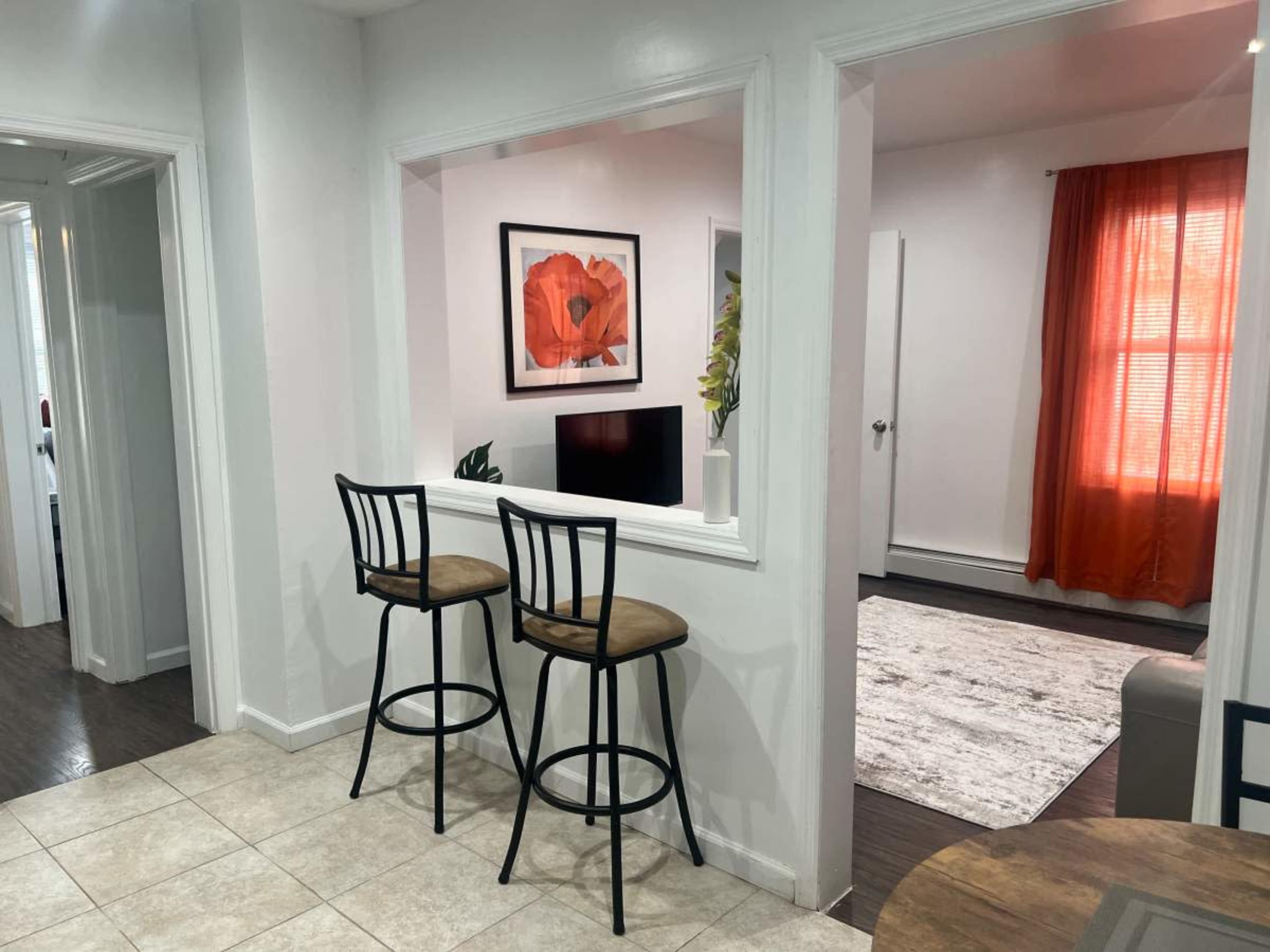 A modern kitchen with two black bar stools facing a narrow opening that leads into a living room adorned with a large floral artwork and a window with orange curtains.