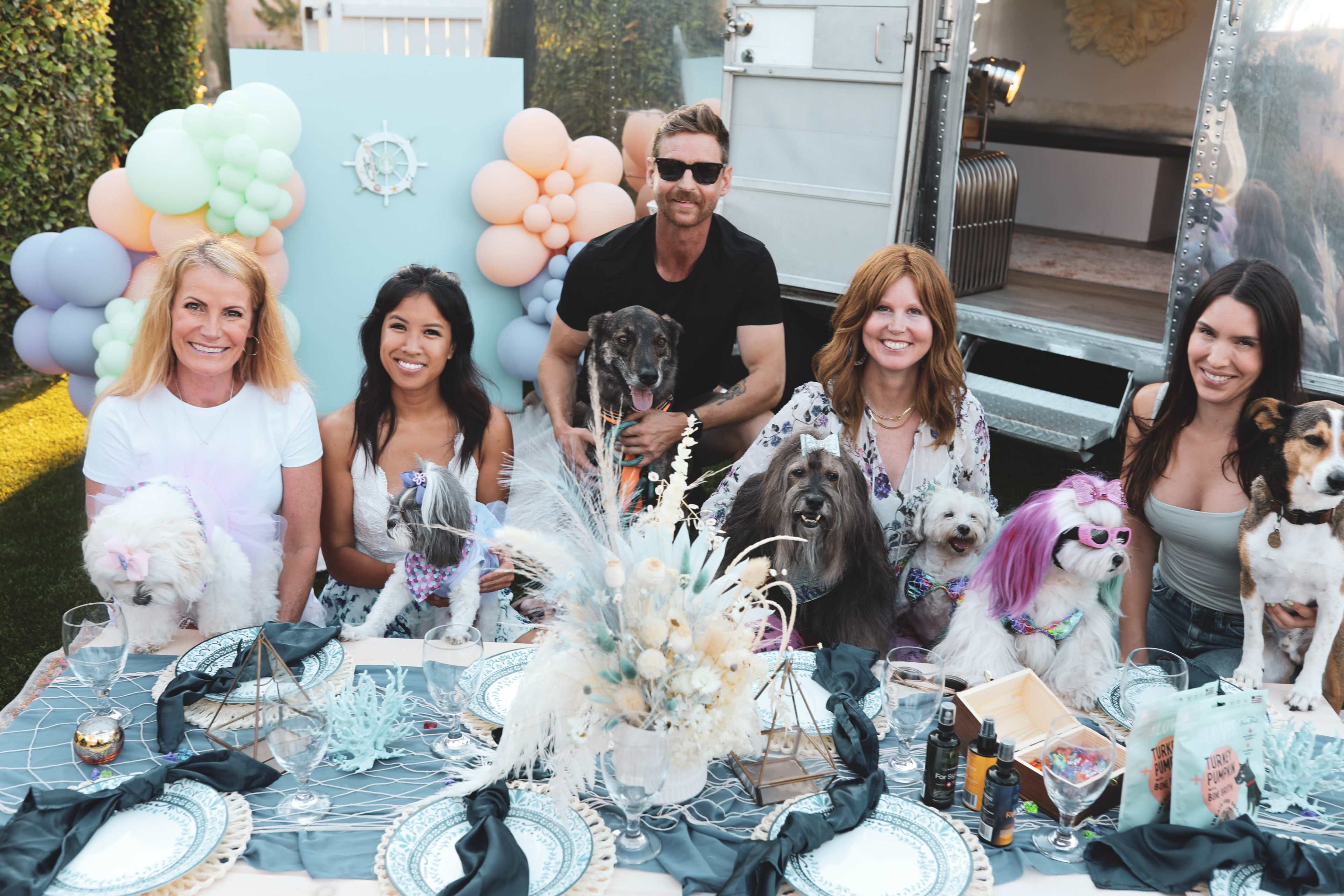 A group of people and several dogs sit around a decorated table outdoors, surrounded by balloons and a vintage trailer.