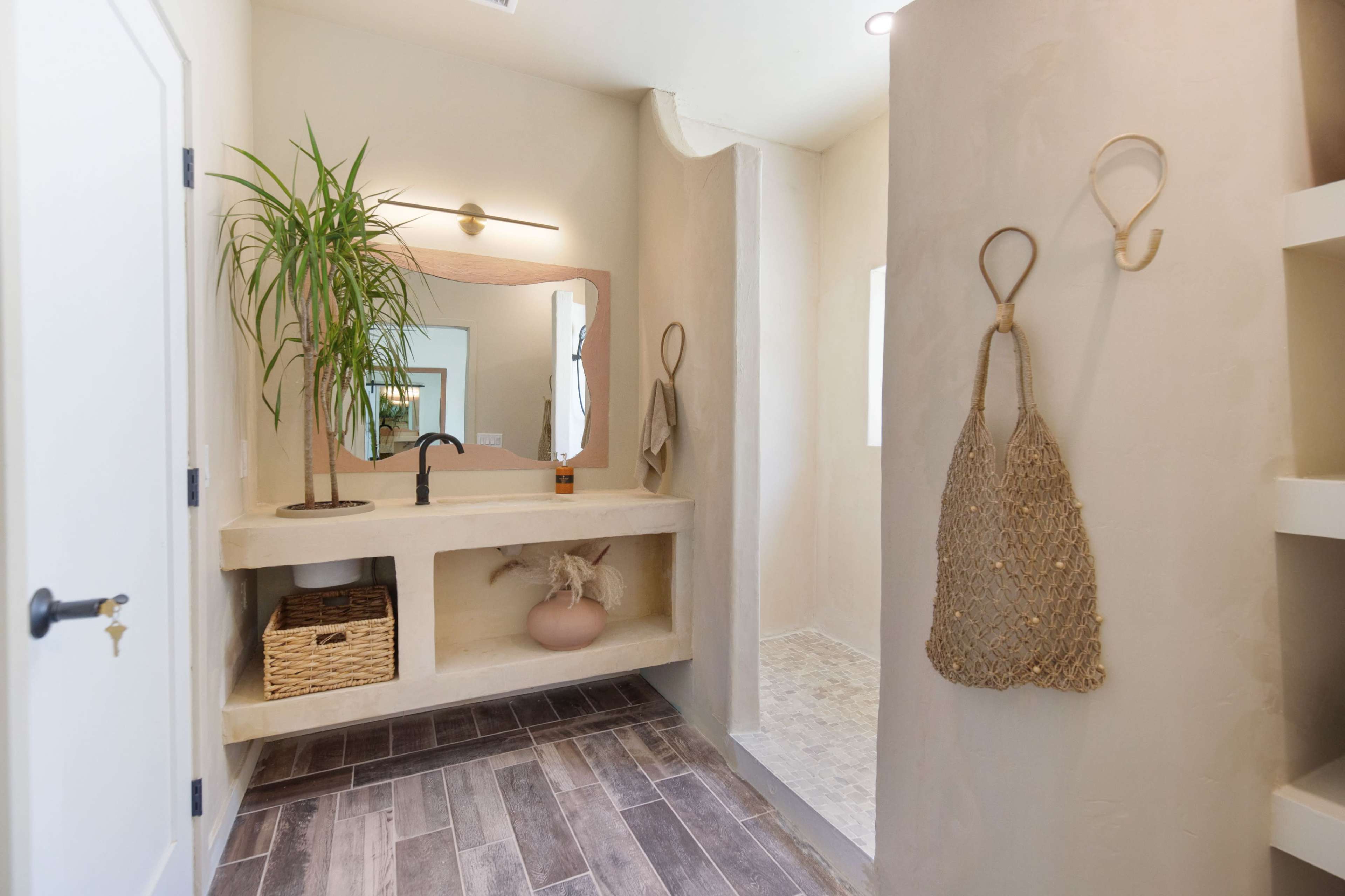 A modern bathroom features a minimalistic design with a plant, a large mirror, and a shower area, complemented by wooden flooring and woven decor.