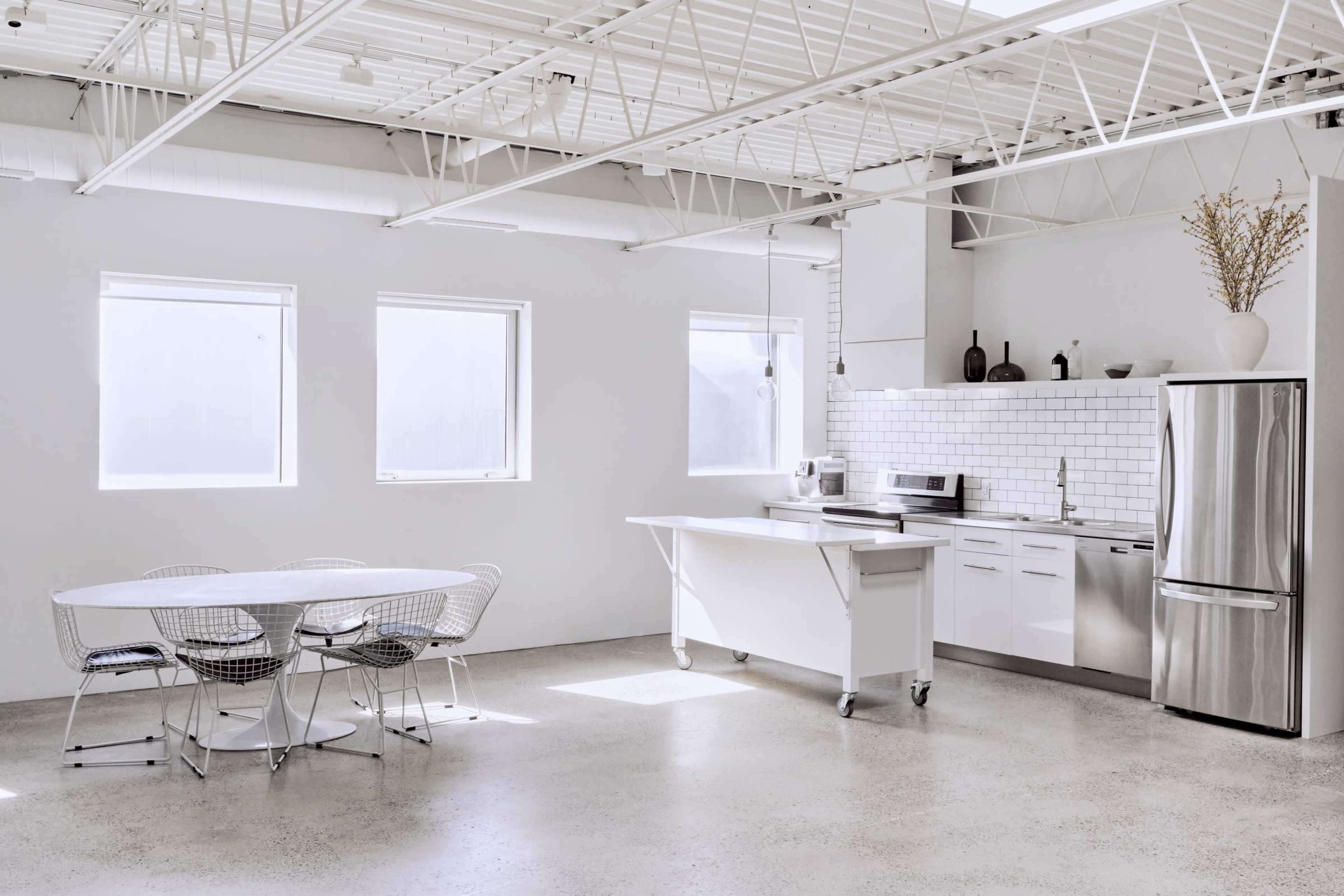 The image shows a modern kitchen and dining area with a minimalist design, featuring a white table and chairs, stainless steel appliances, and large windows that let in natural light.