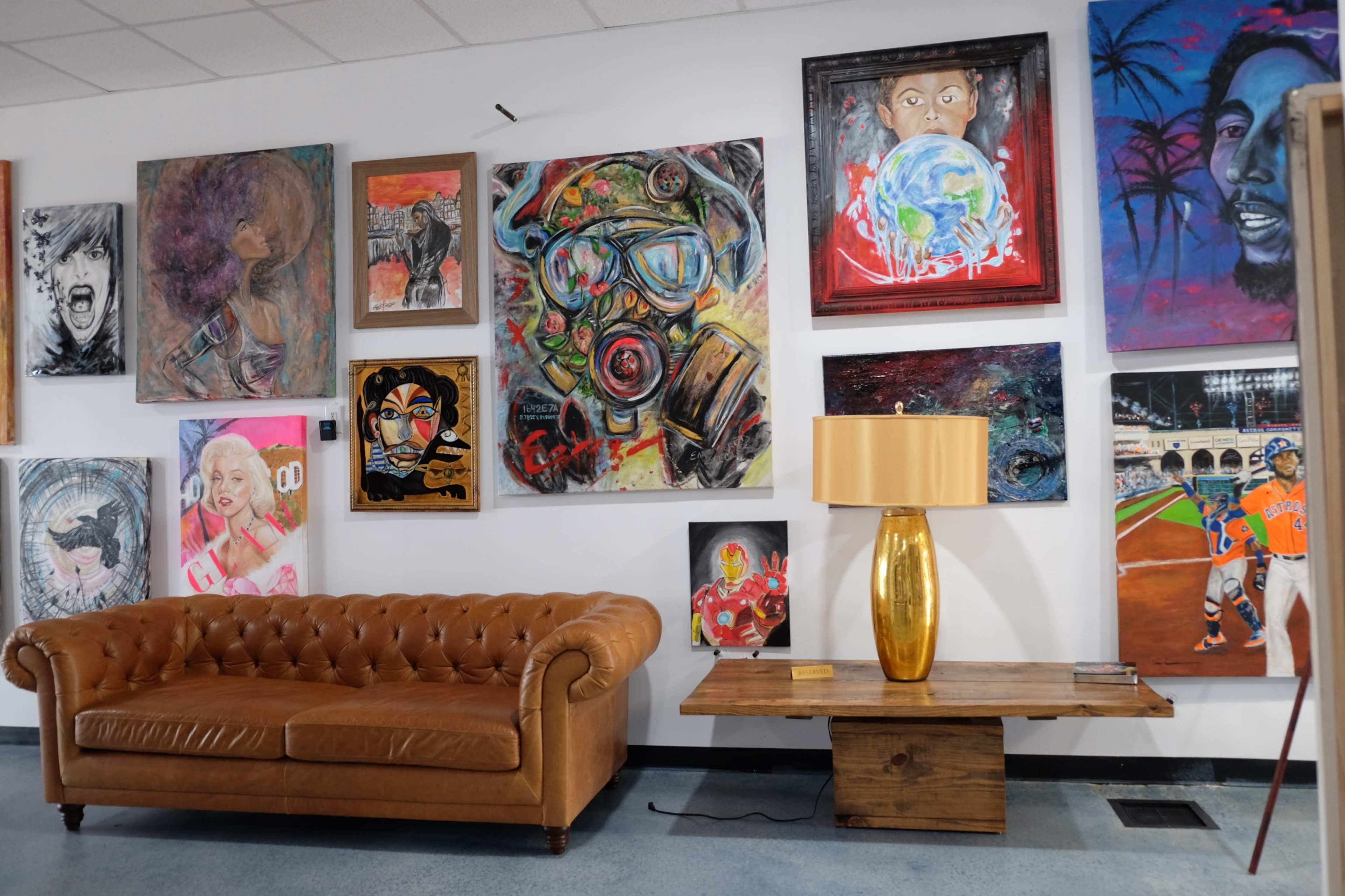 A leather sofa in front of a gallery-style wall adorned with various colorful paintings and a wooden table with a gold lamp.