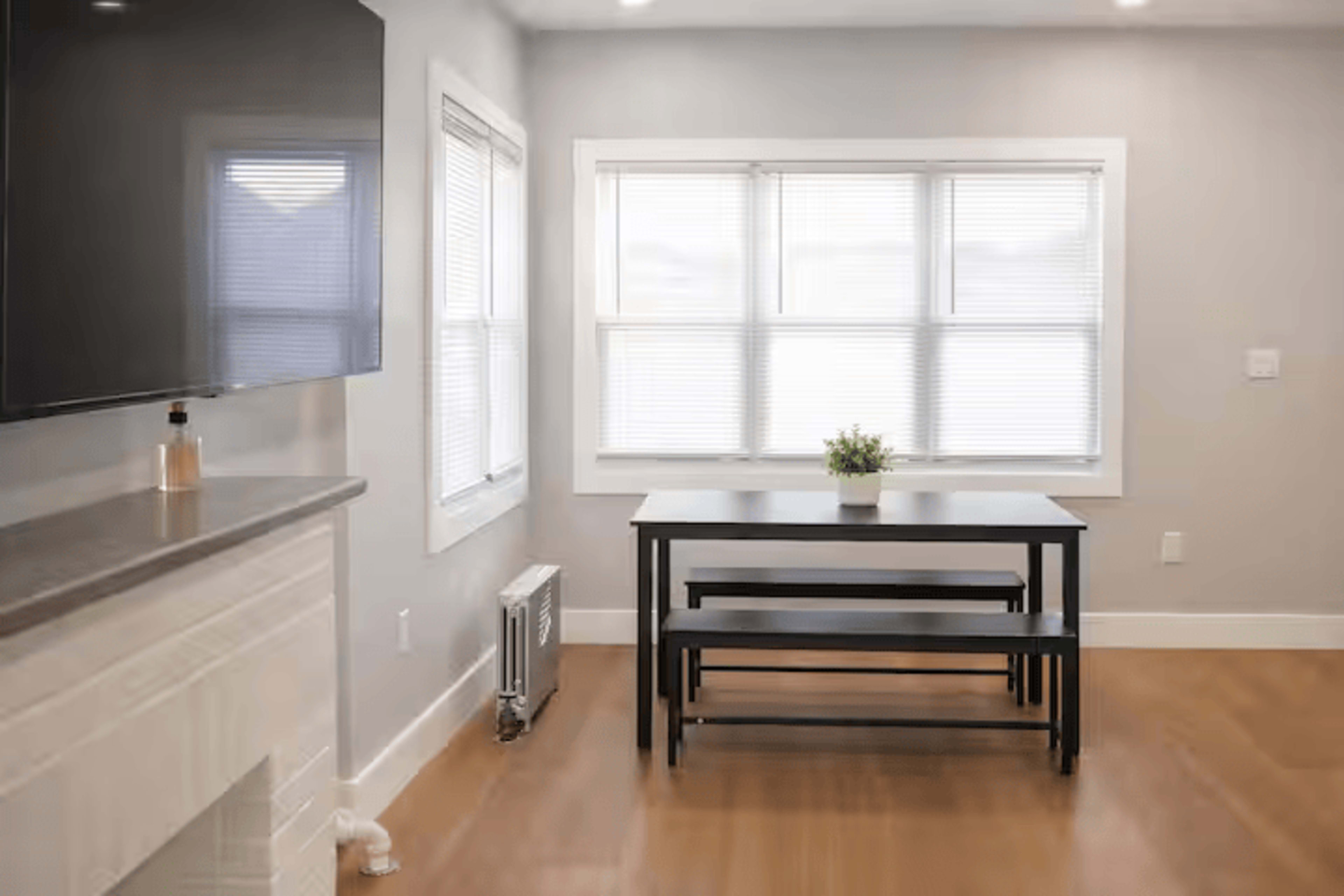 A simple dining area features a black table and benches, alongside large windows with blinds, and a gray wall with a television mounted nearby.