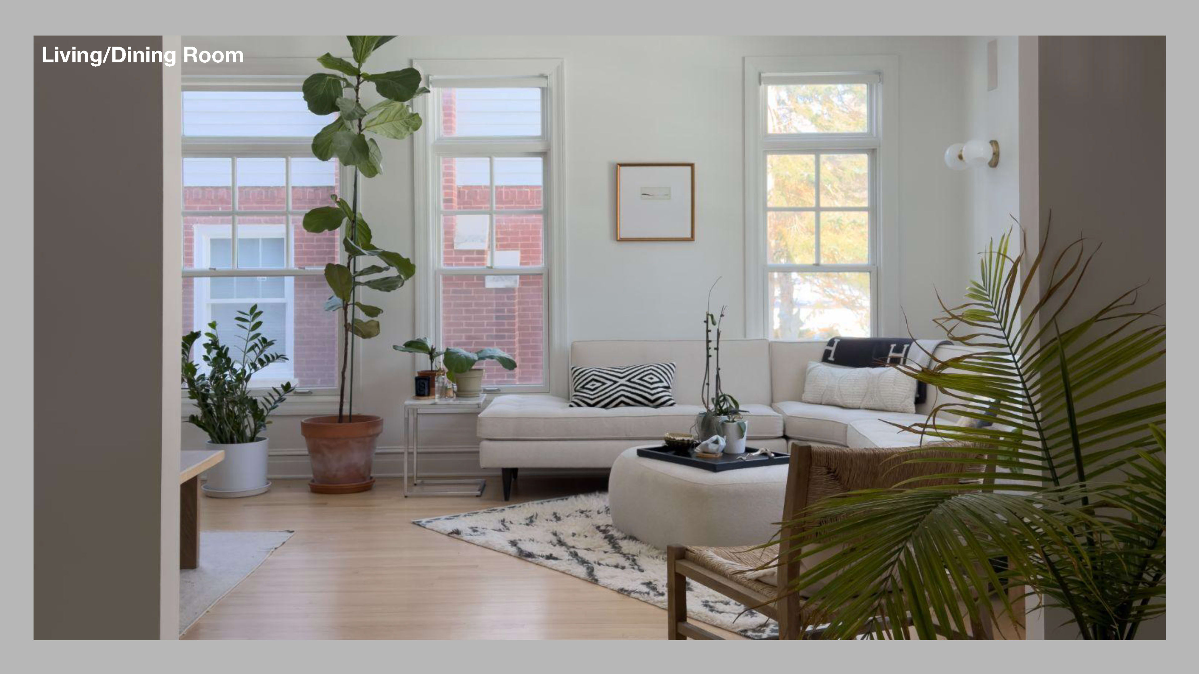 The image shows a modern living and dining room featuring large windows, a light-colored sectional sofa, a round coffee table, and various indoor plants.