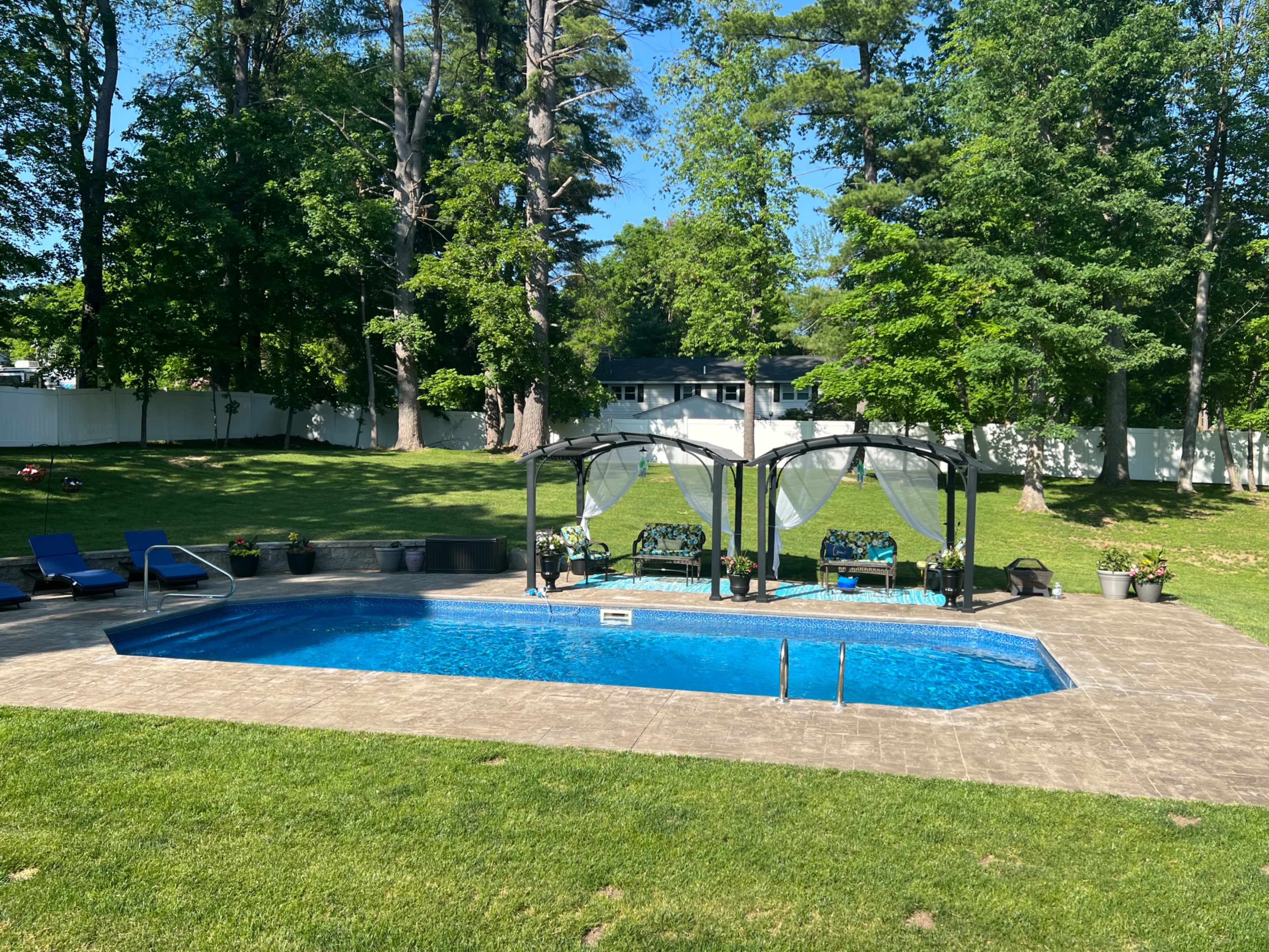 The image shows a clear swimming pool surrounded by a well-maintained lawn, with lounging chairs and shaded seating areas in a backyard setting.