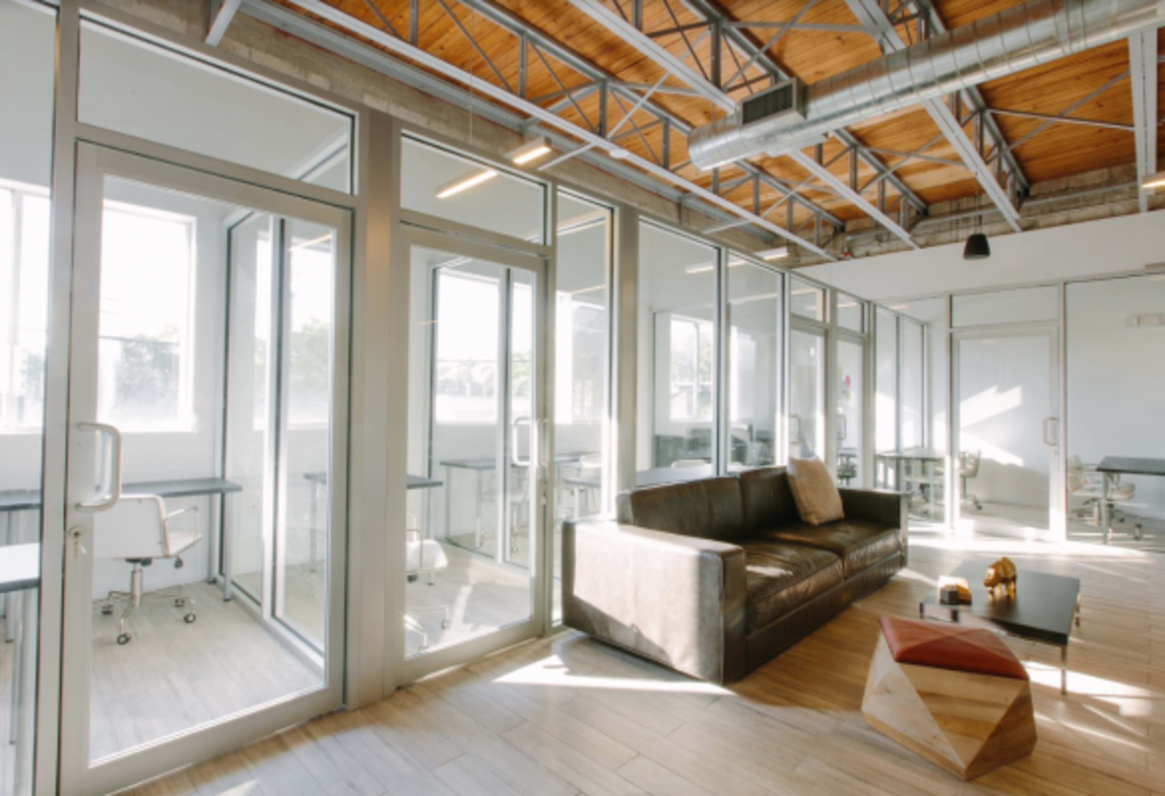 The image shows a modern office space with large glass walls, a brown sofa, and several workstations in a well-lit environment.
