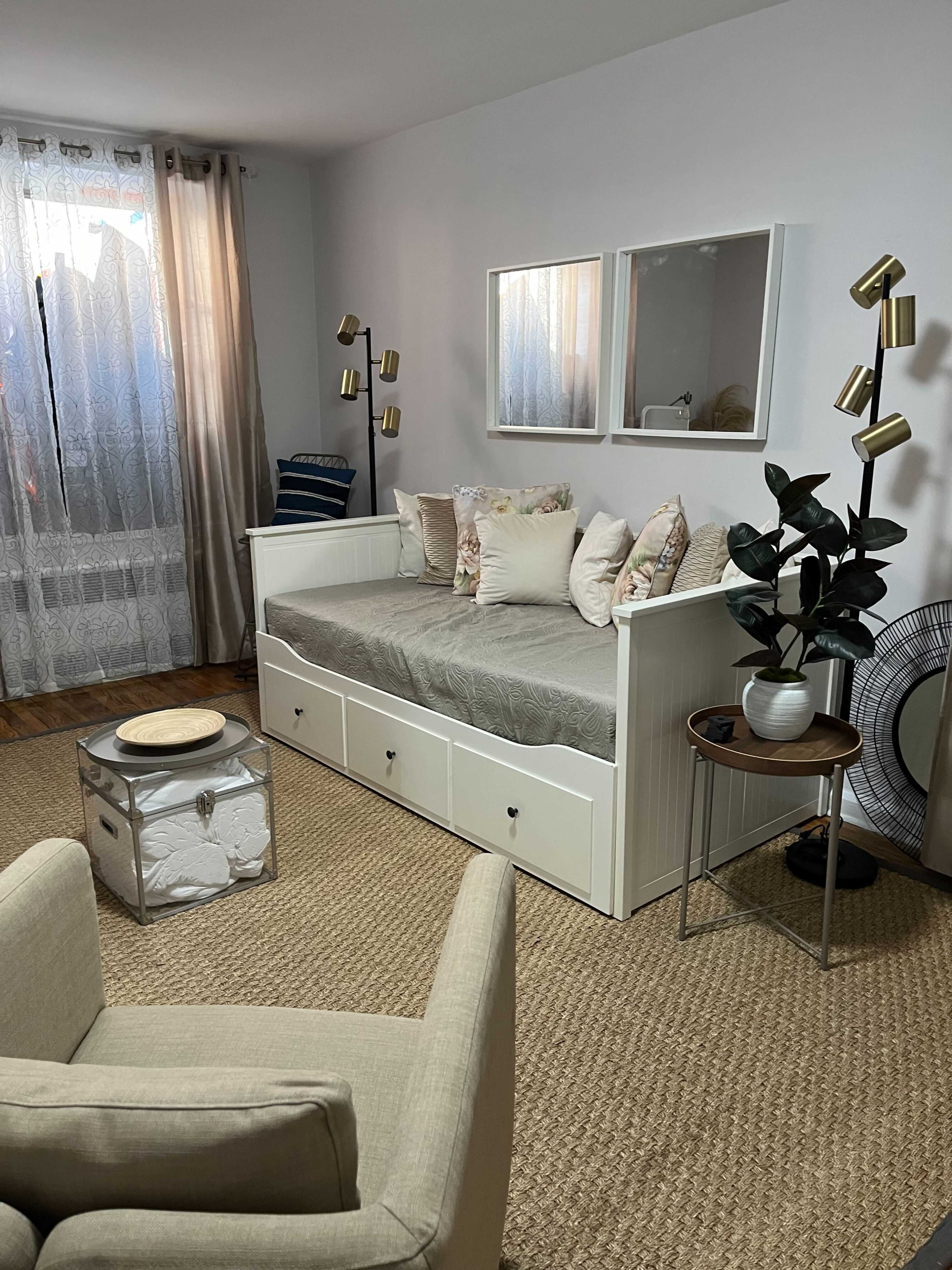 The image shows a cozy room featuring a daybed with decorative pillows, a small side table, a plant, and a neutral-colored armchair.