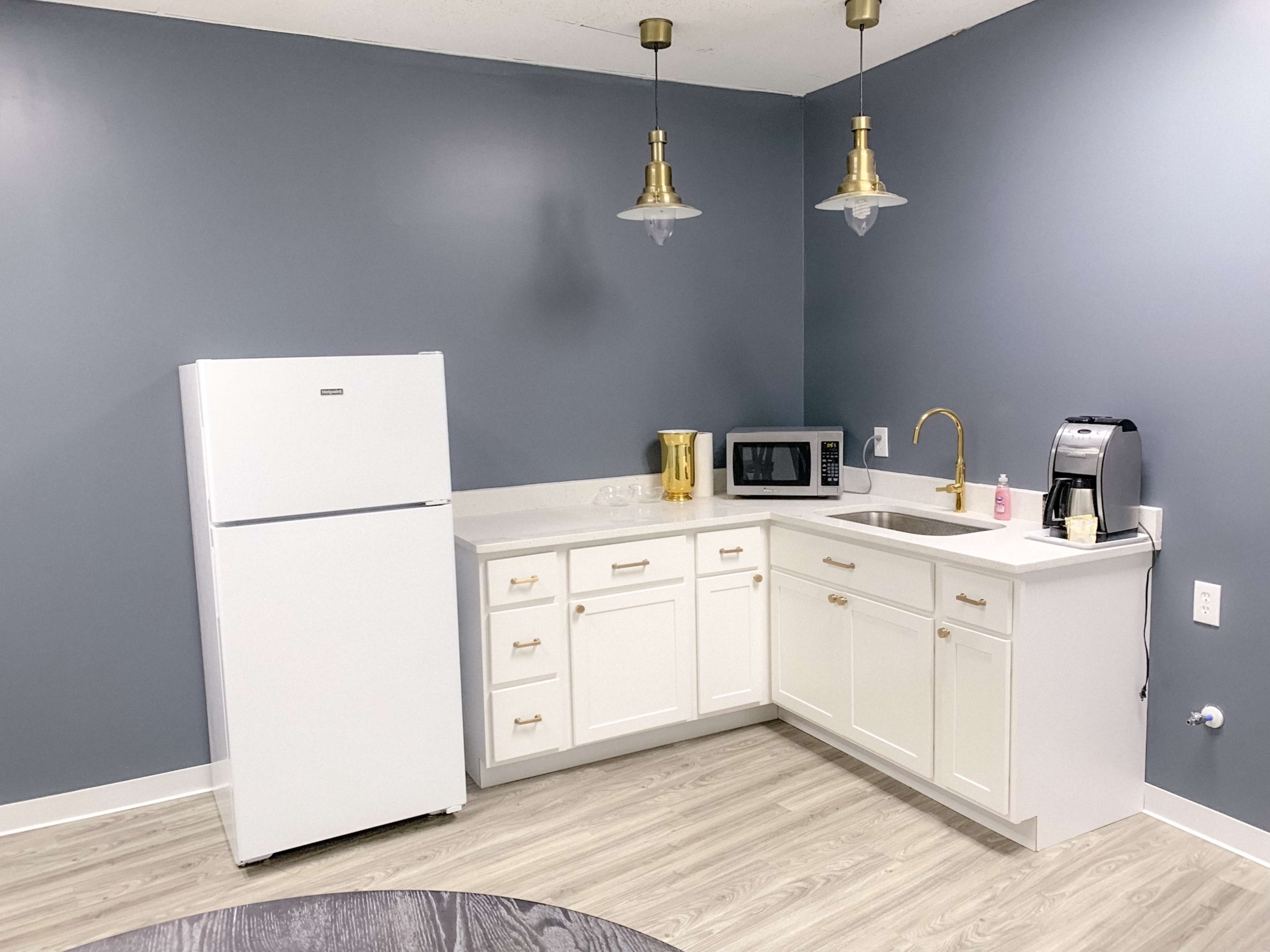 A modern kitchen with white cabinetry, a refrigerator, sink, microwave, and coffee maker against a gray wall.