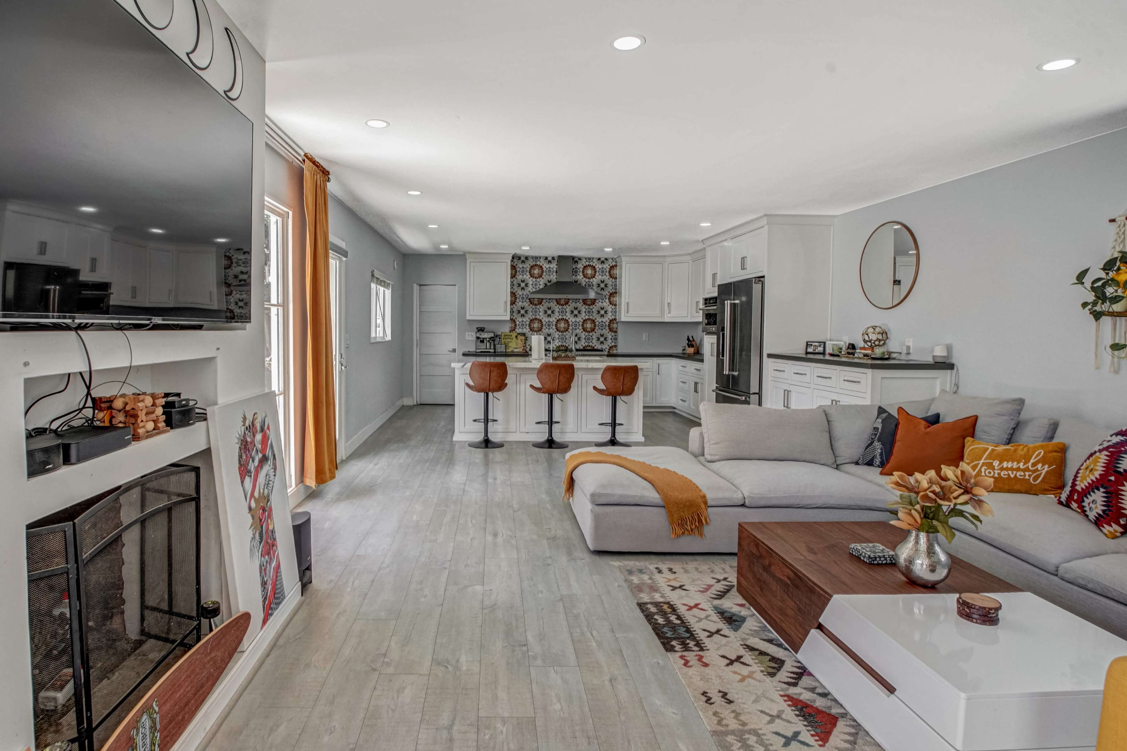 A modern living room features a large sectional sofa, a coffee table, a flat-screen TV, and an open kitchen with bar stools against a patterned backsplash.