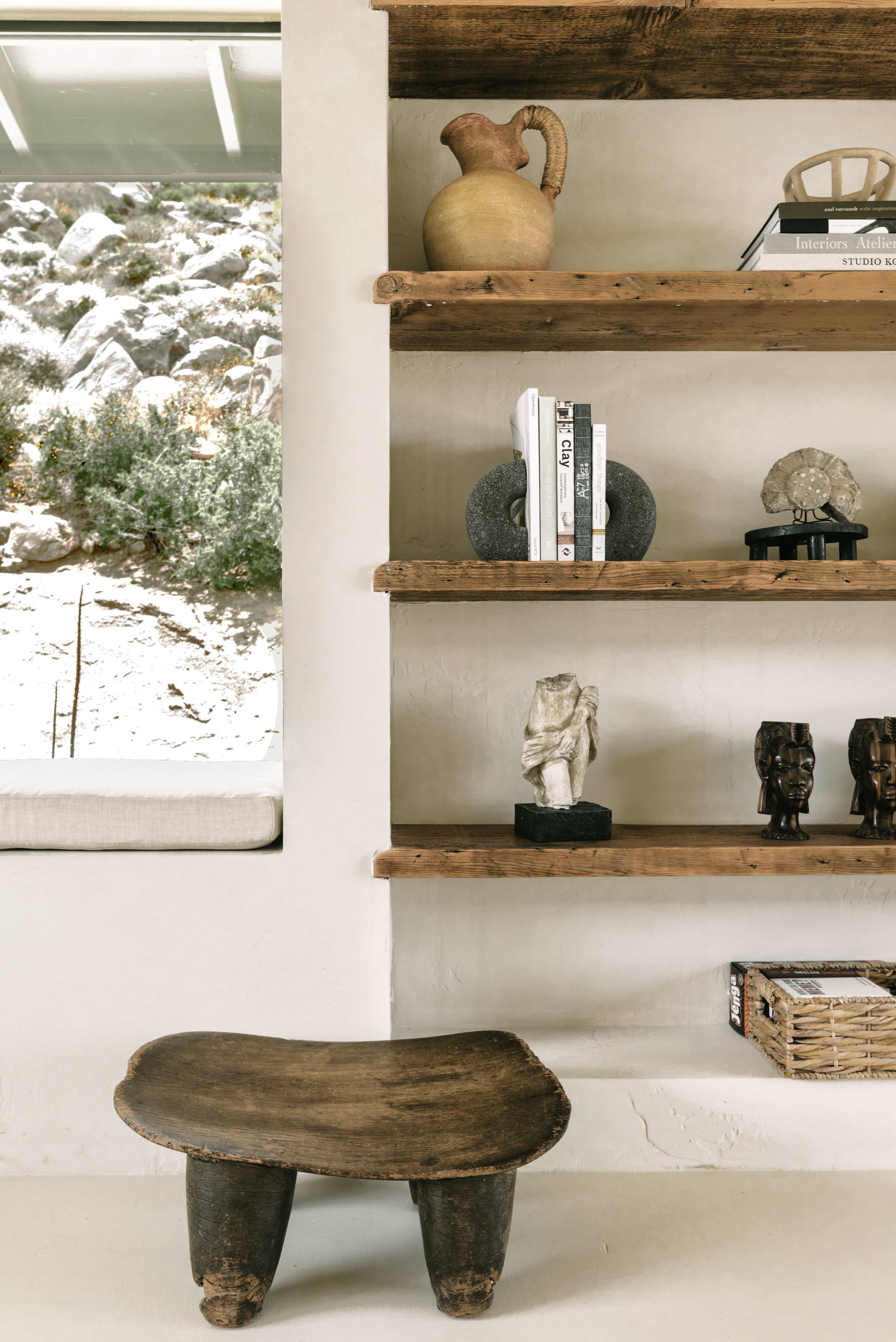 The image features a wooden stool in front of a shelf displaying various decorative objects and books, alongside a large window revealing a rocky landscape outside.