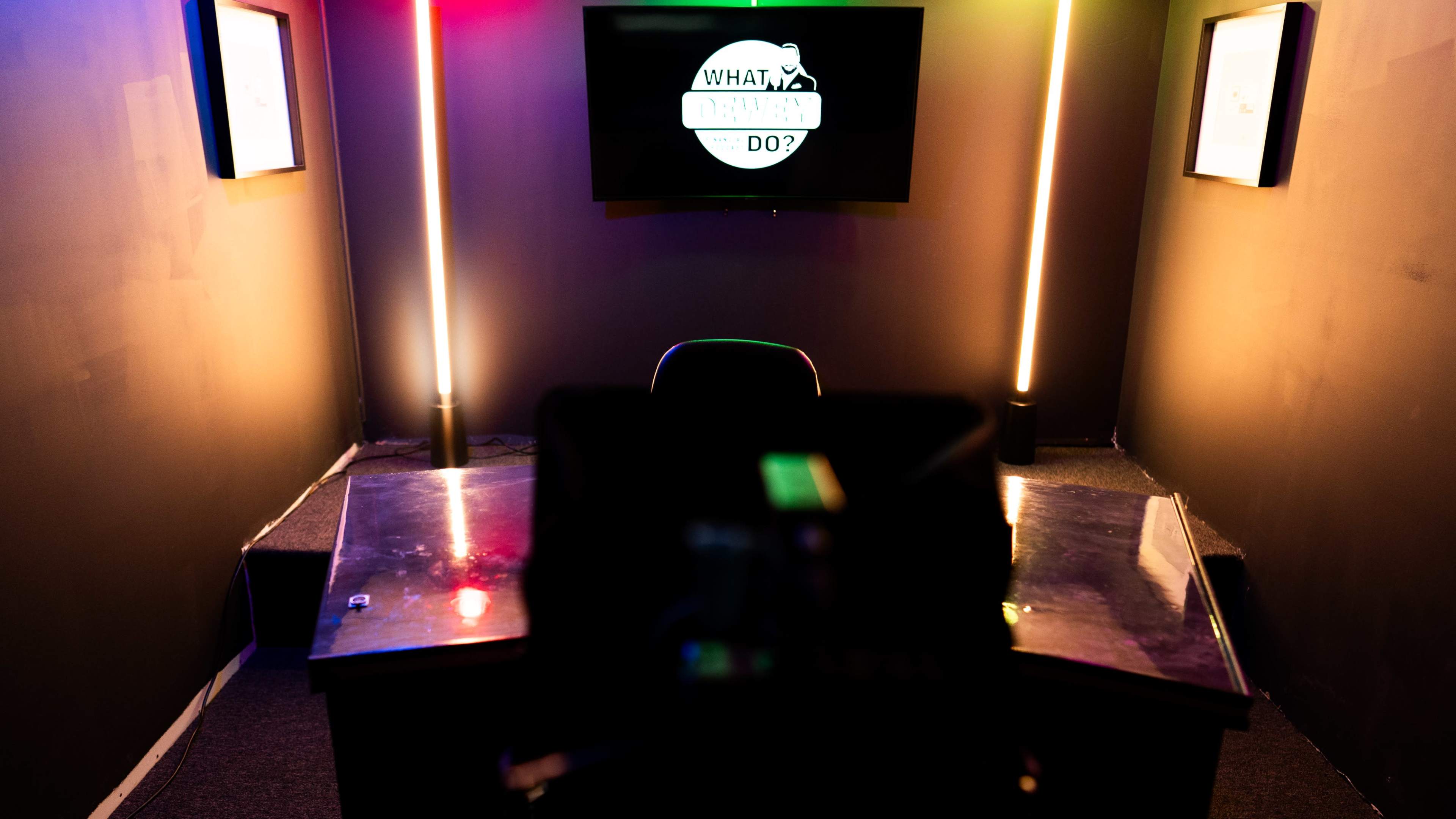 A dimly lit studio features a central desk with a chair facing a screen displaying "What Would You Do?" on the wall.