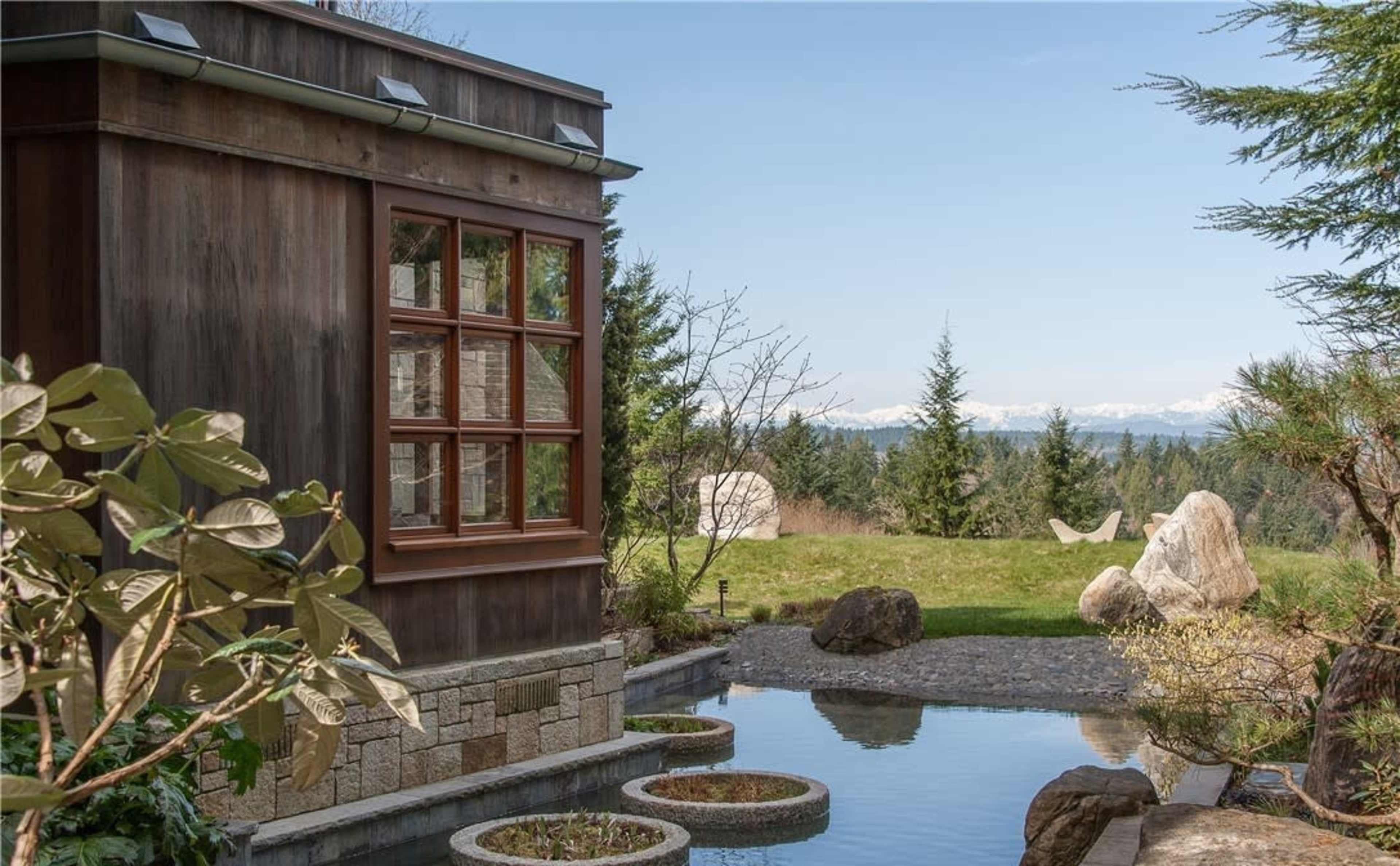 A wooden structure with large windows overlooks a landscaped garden featuring a pond, rocks, and distant mountains.