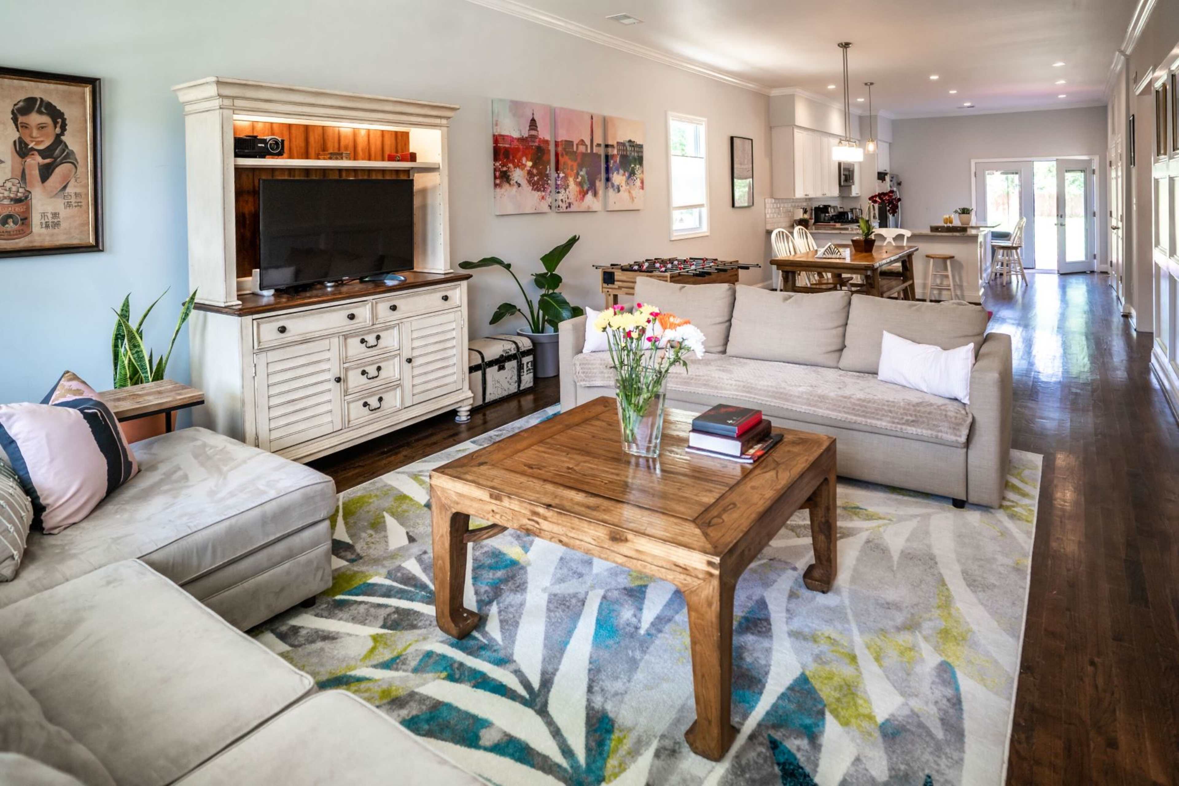 A spacious living room with a light-colored sectional sofa, a wooden coffee table, a television on a white cabinet, and a view of an open kitchen in the background.
