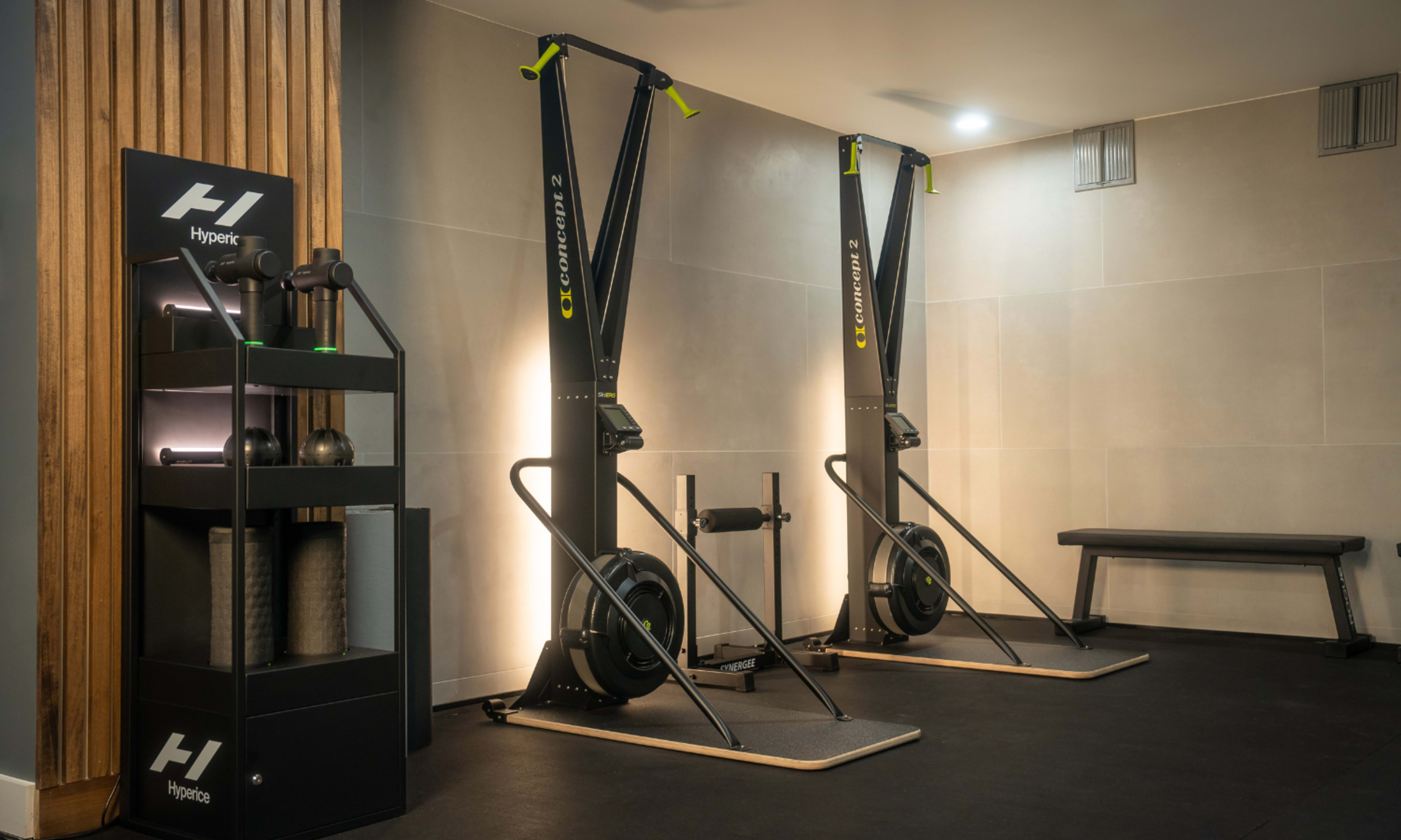 A modern gym area featuring two rowing machines and a storage rack with fitness equipment against a minimalist wall.