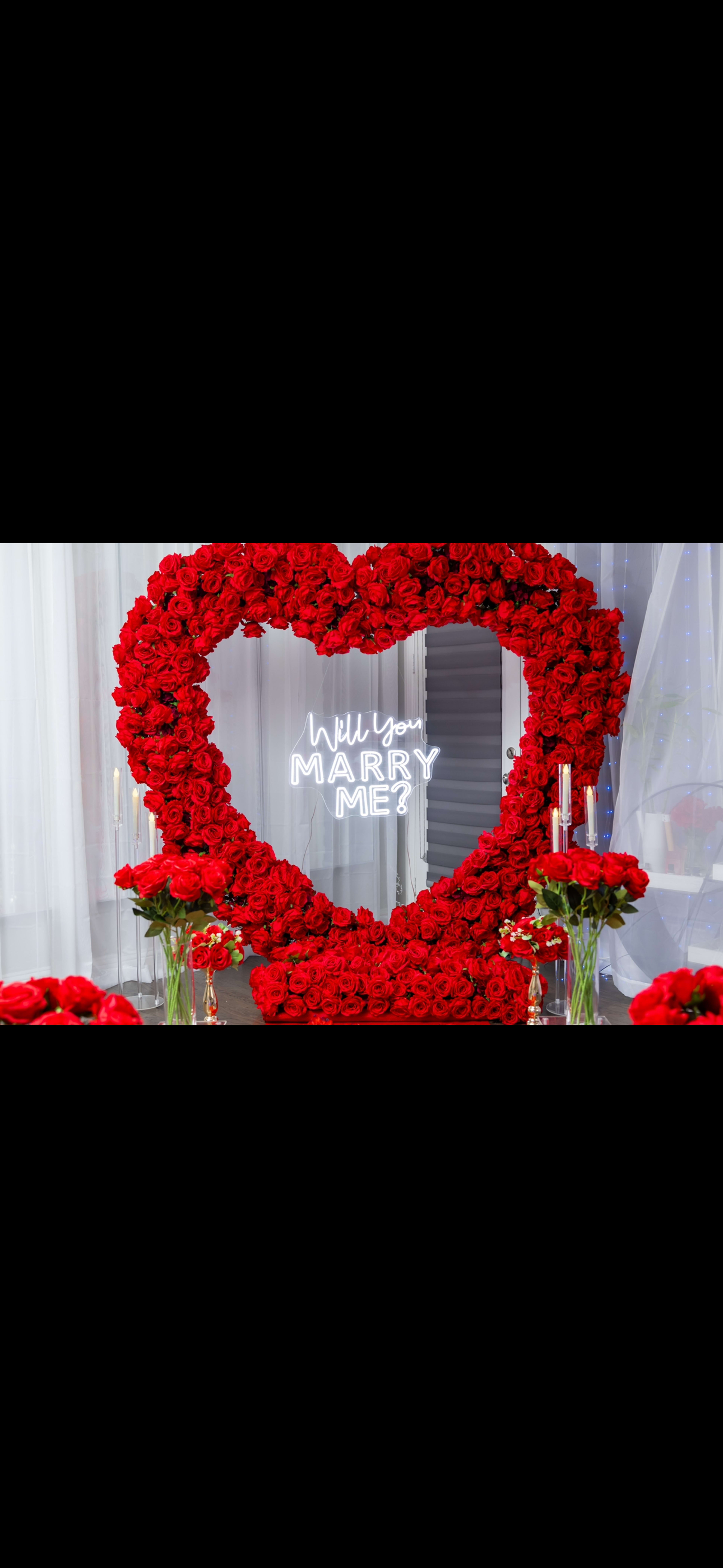 A large heart-shaped arrangement of red roses surrounds a neon sign that reads "Will You Marry Me?" in a decorated room.
