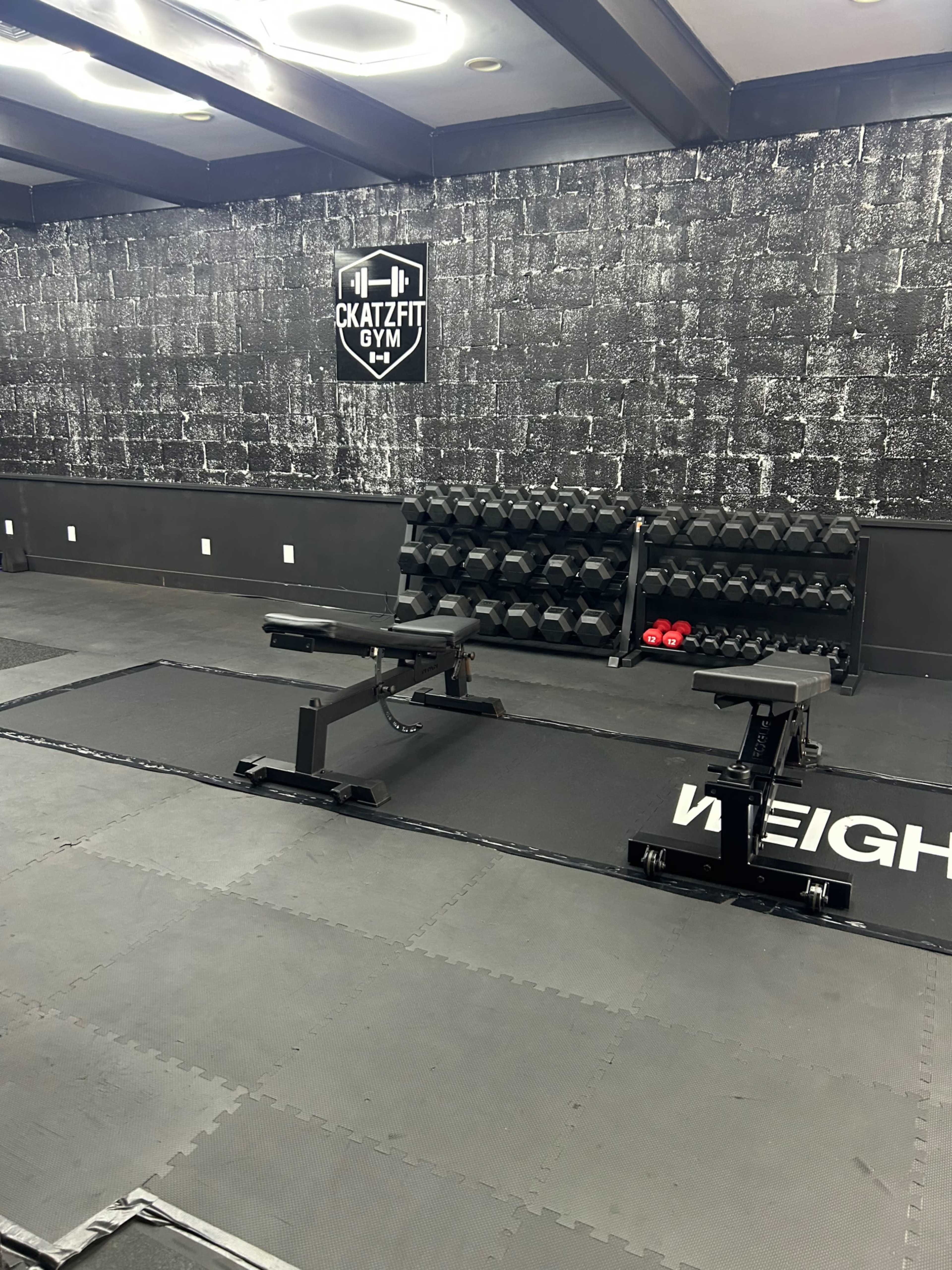 The image shows a gym interior with a black and white theme, featuring dumbbells, a bench, and exercise mats on the floor.