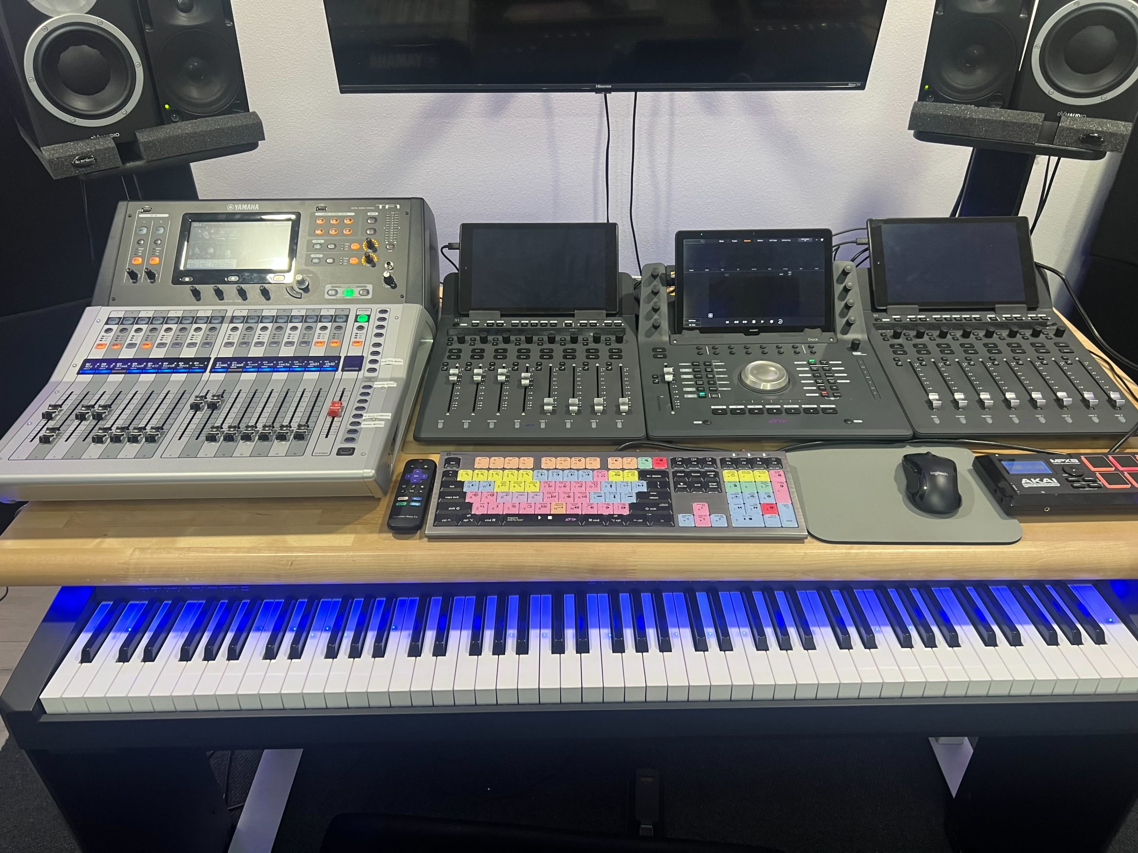 A digital audio workstation setup featuring a mixing console, multiple touchscreen controllers, and a MIDI keyboard, all arranged on a desk with LED lighting.