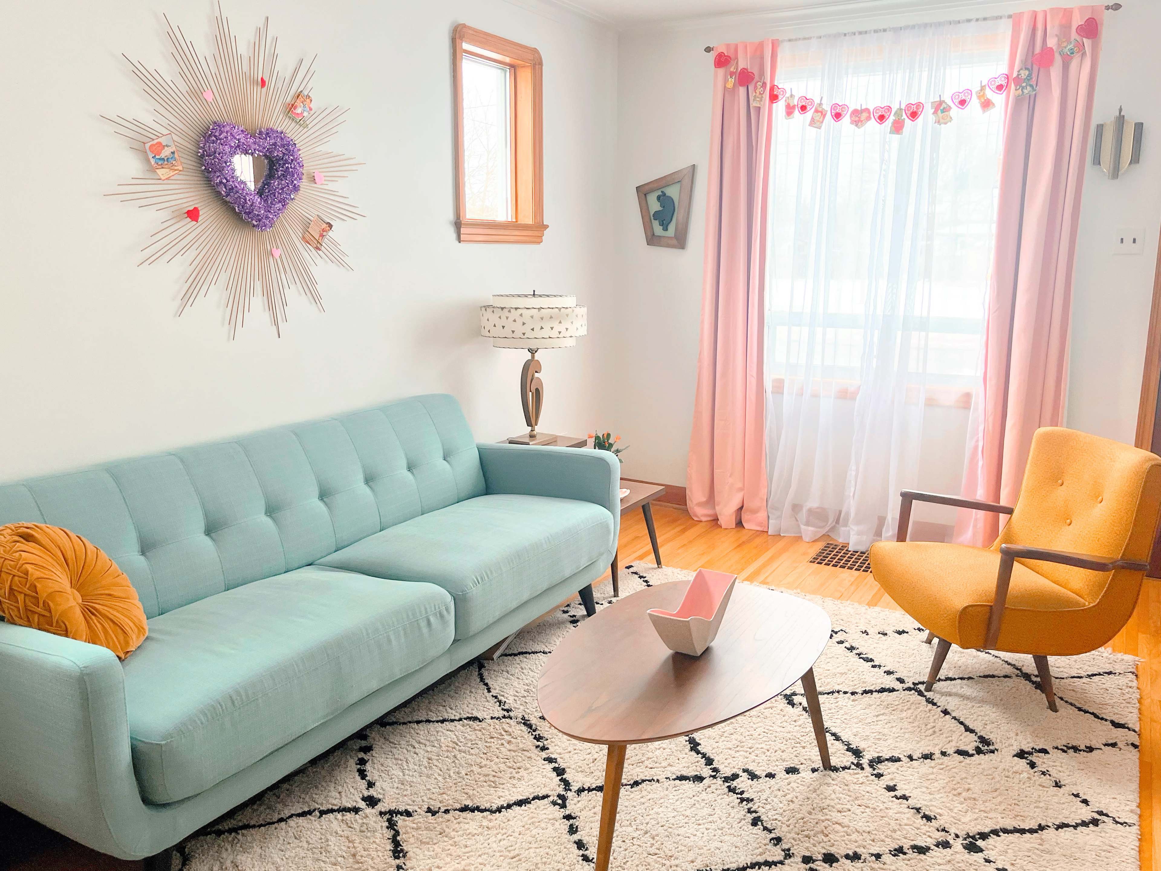 A living room featuring a light blue sofa, an orange armchair, a wooden table, and bright curtains beside a window.
