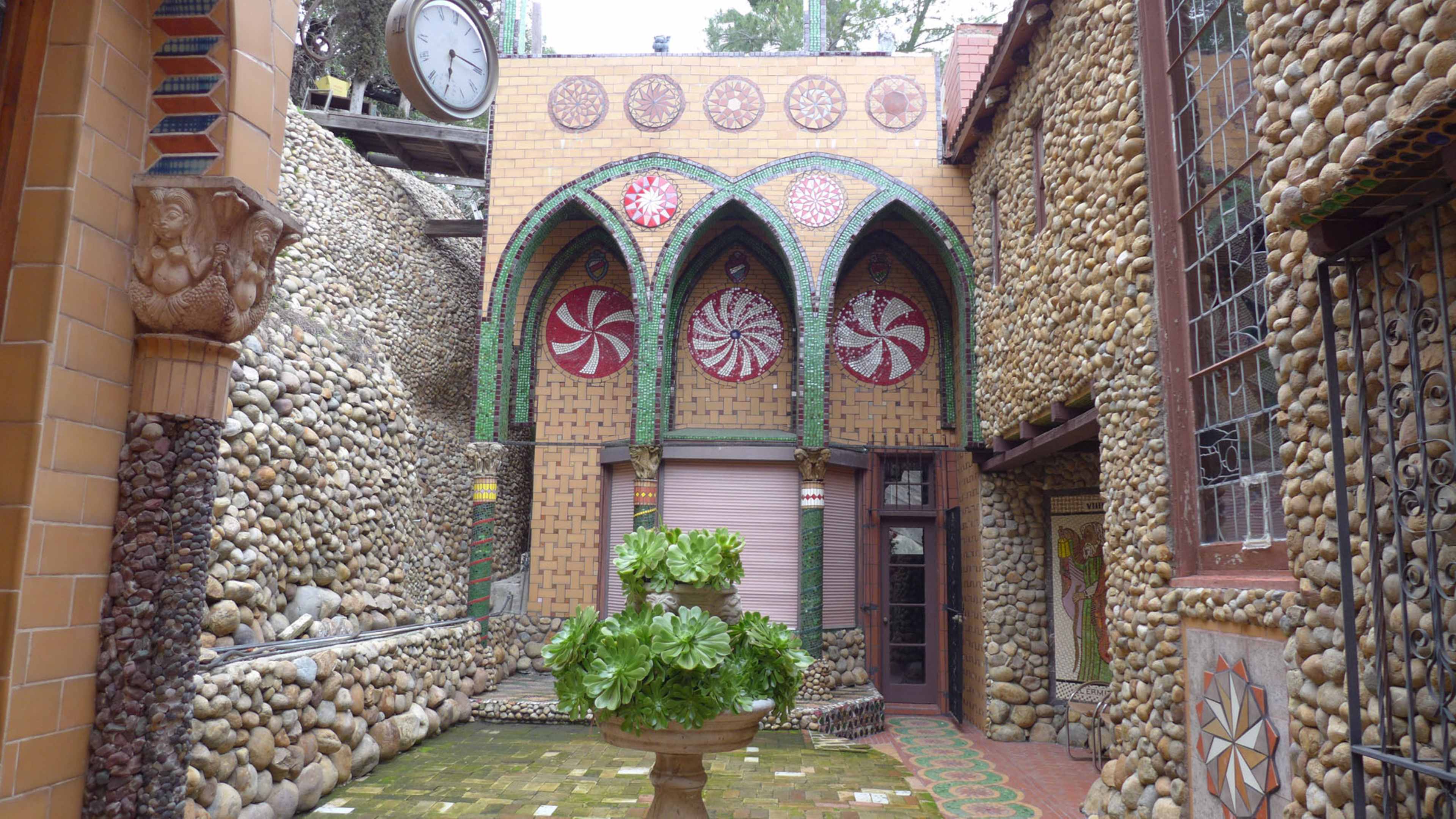 The image features a courtyard with stone walls, arched decorative elements, and a central planter surrounded by patterned tiles.