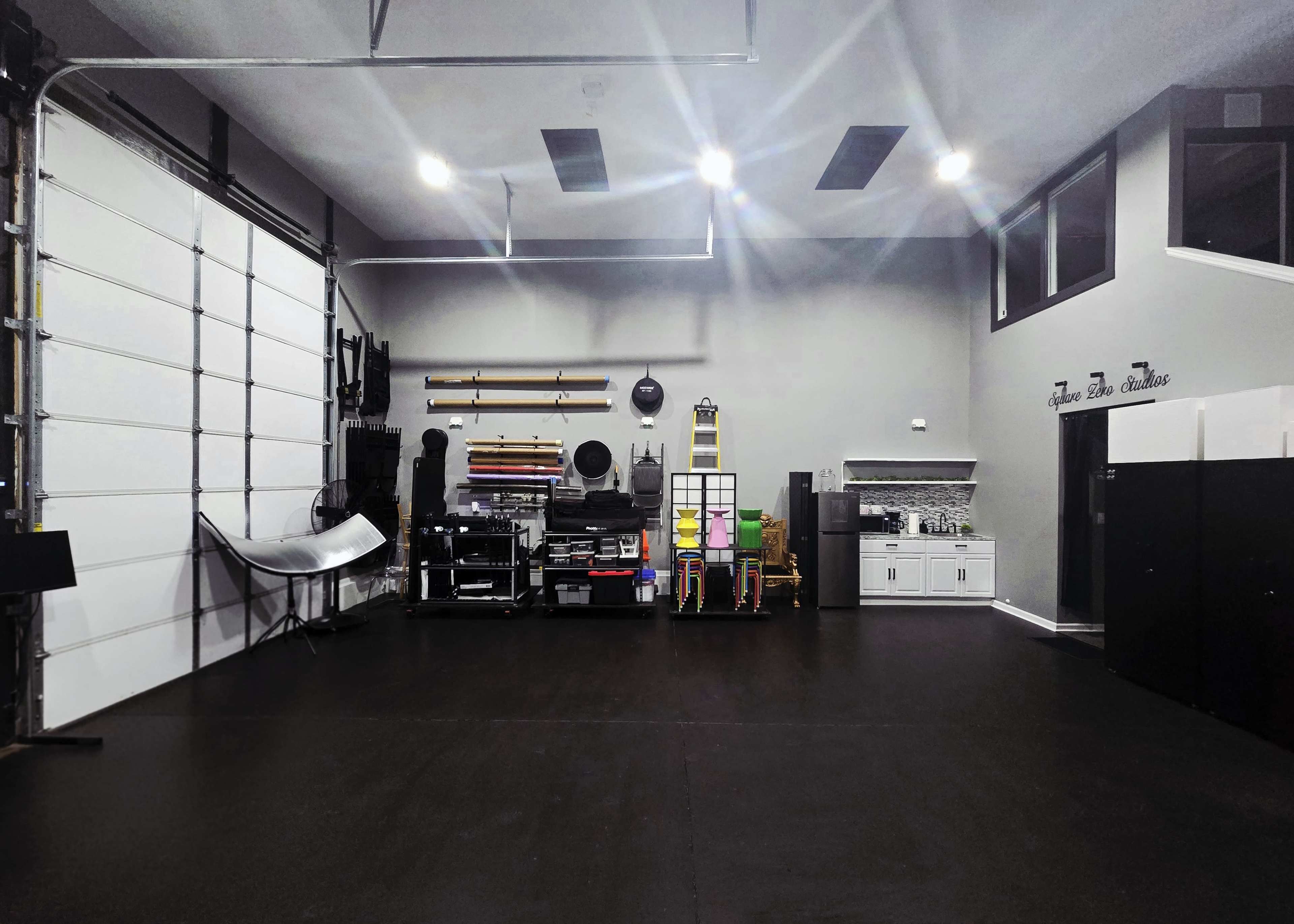 The image shows a spacious garage with a large white sectional door, organized shelves, various tools, and a kitchenette area along one wall.