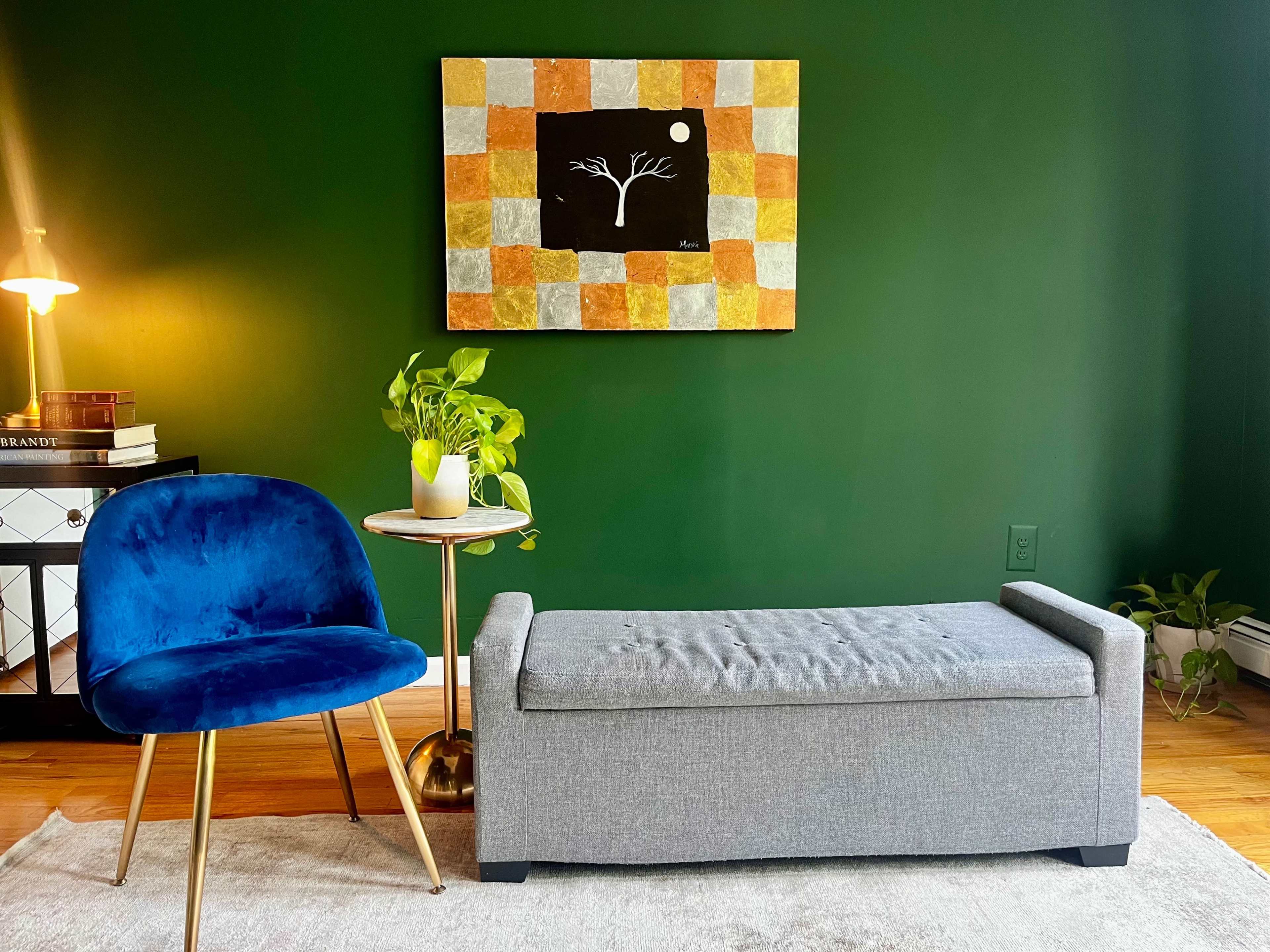 The scene features a gray upholstered bench, a blue velvet chair, a round side table with a plant, and a colorful abstract painting on a dark green wall.