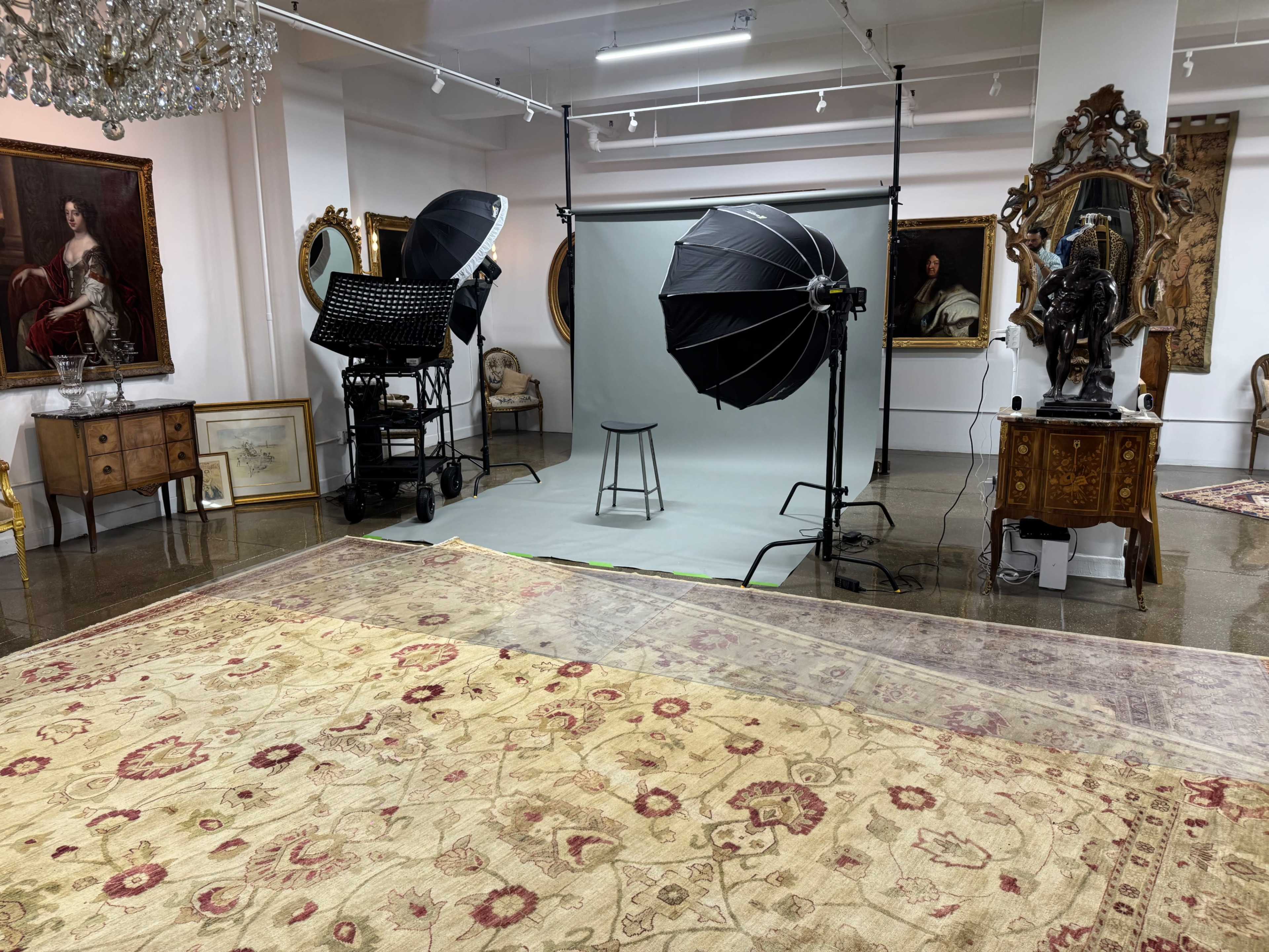 The image shows a photography studio setup featuring two large softbox lights, a stool in front of a green backdrop, and several antique furnishings and artworks around the space.