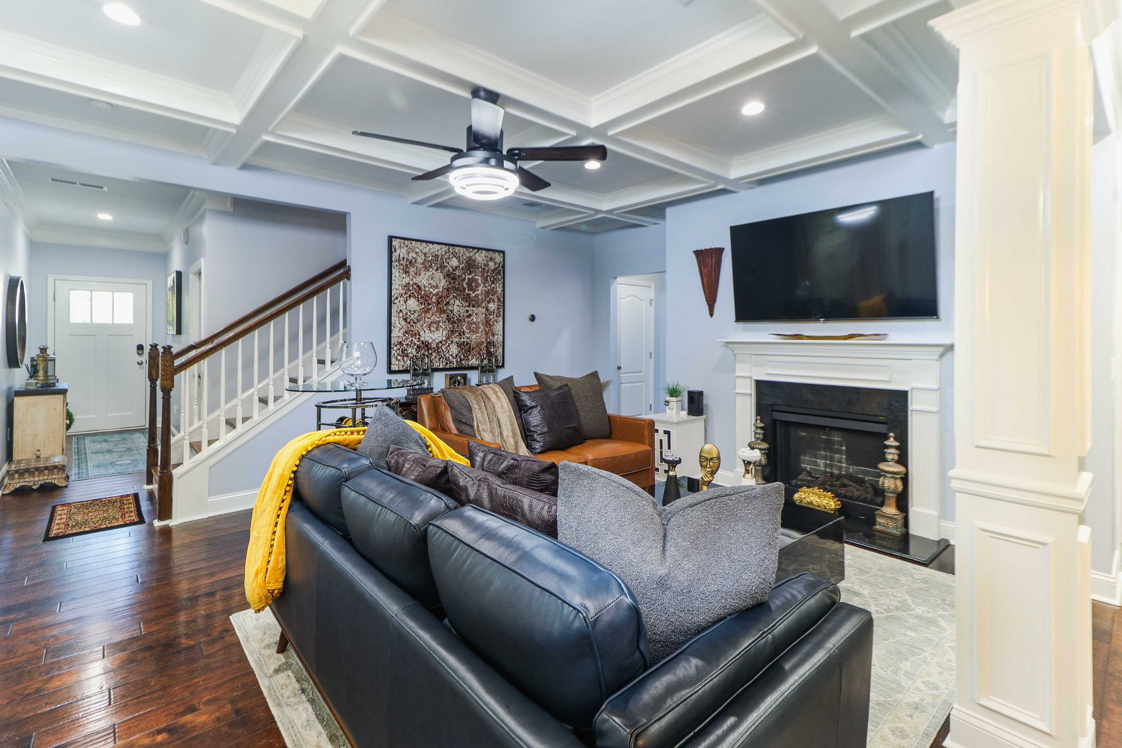 The living room features a dark leather sofa, a large flat-screen TV mounted above a white fireplace, and a staircase leading to another level.