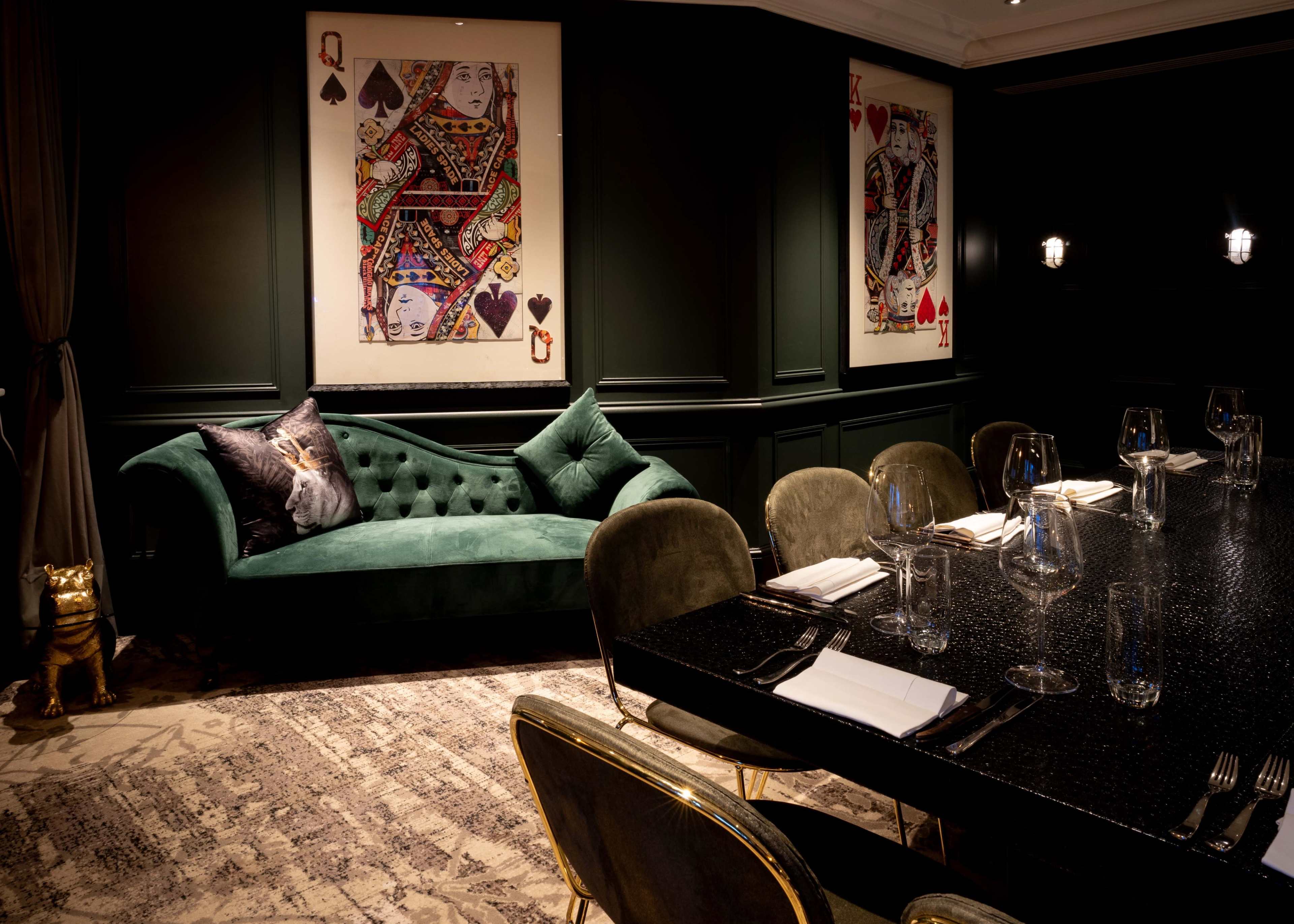 The image depicts an elegantly decorated dining room featuring a dark green velvet couch, a long black dining table set with glassware, and framed playing card artwork on the walls.