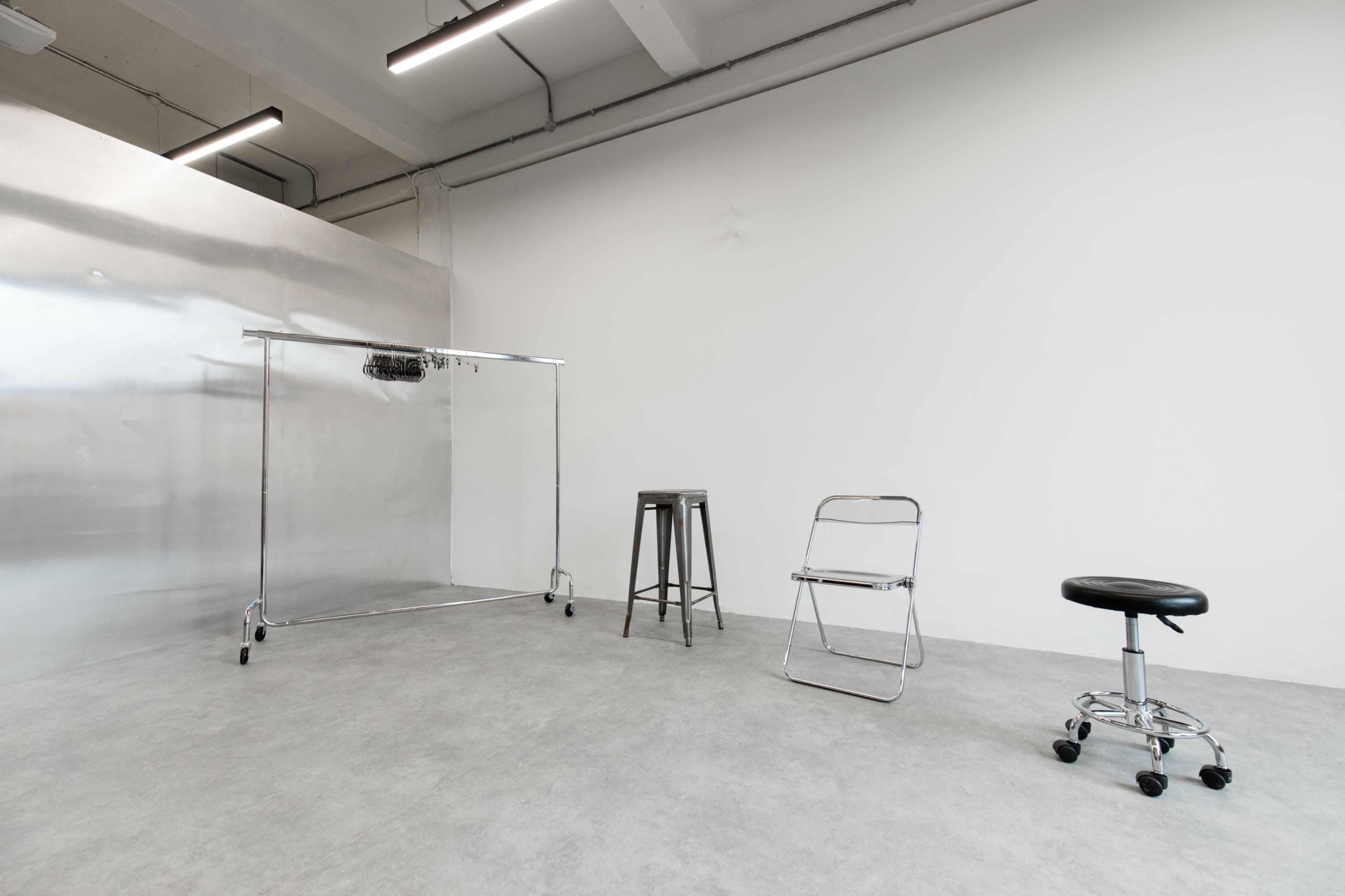 The image shows an empty room with a silver clothing rack, a metal stool, a folding chair, and a rolling stool on a concrete floor.