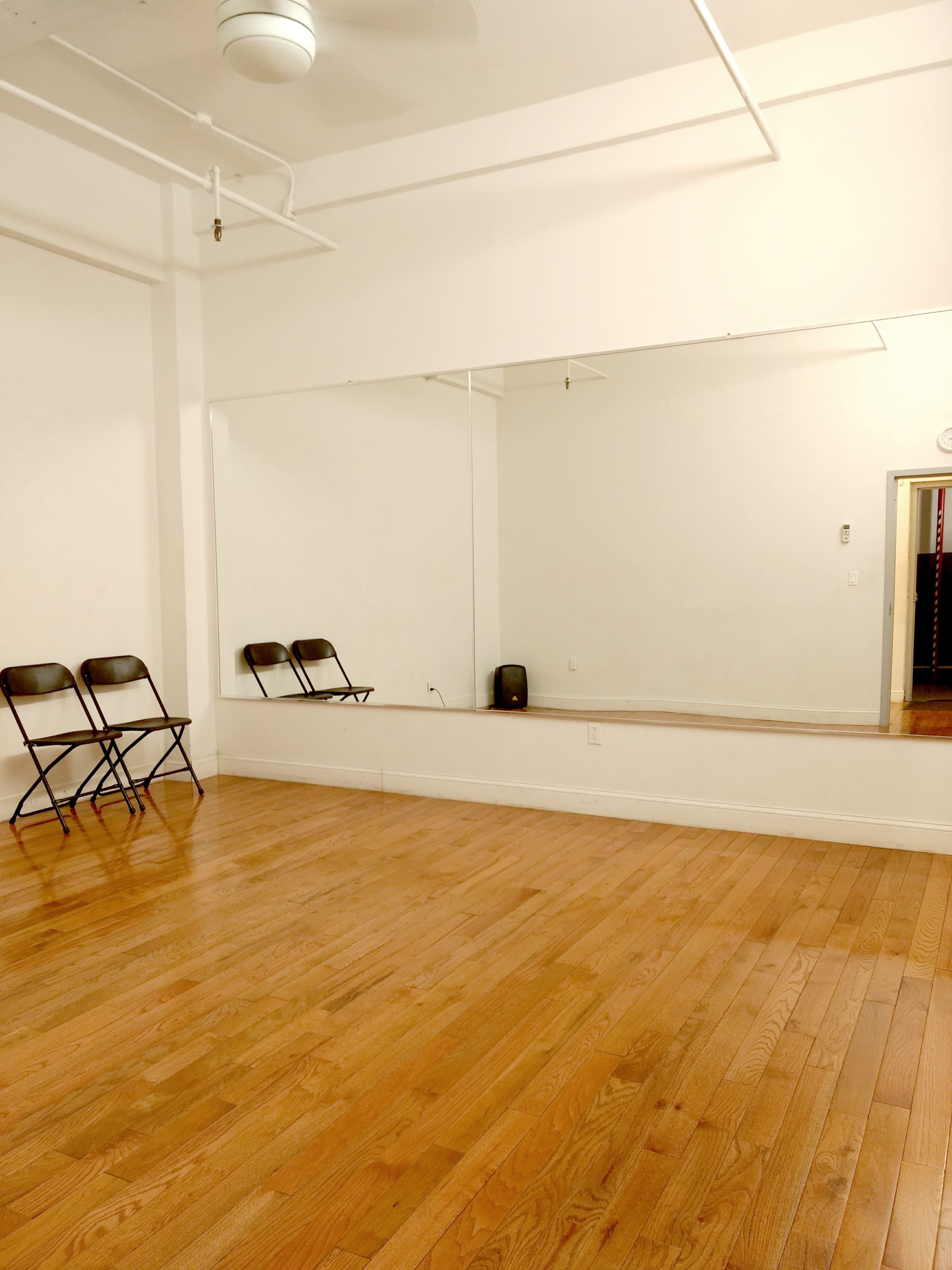 A spacious room with wooden flooring, three black folding chairs lined up against a mirrored wall, and a ceiling fan overhead.