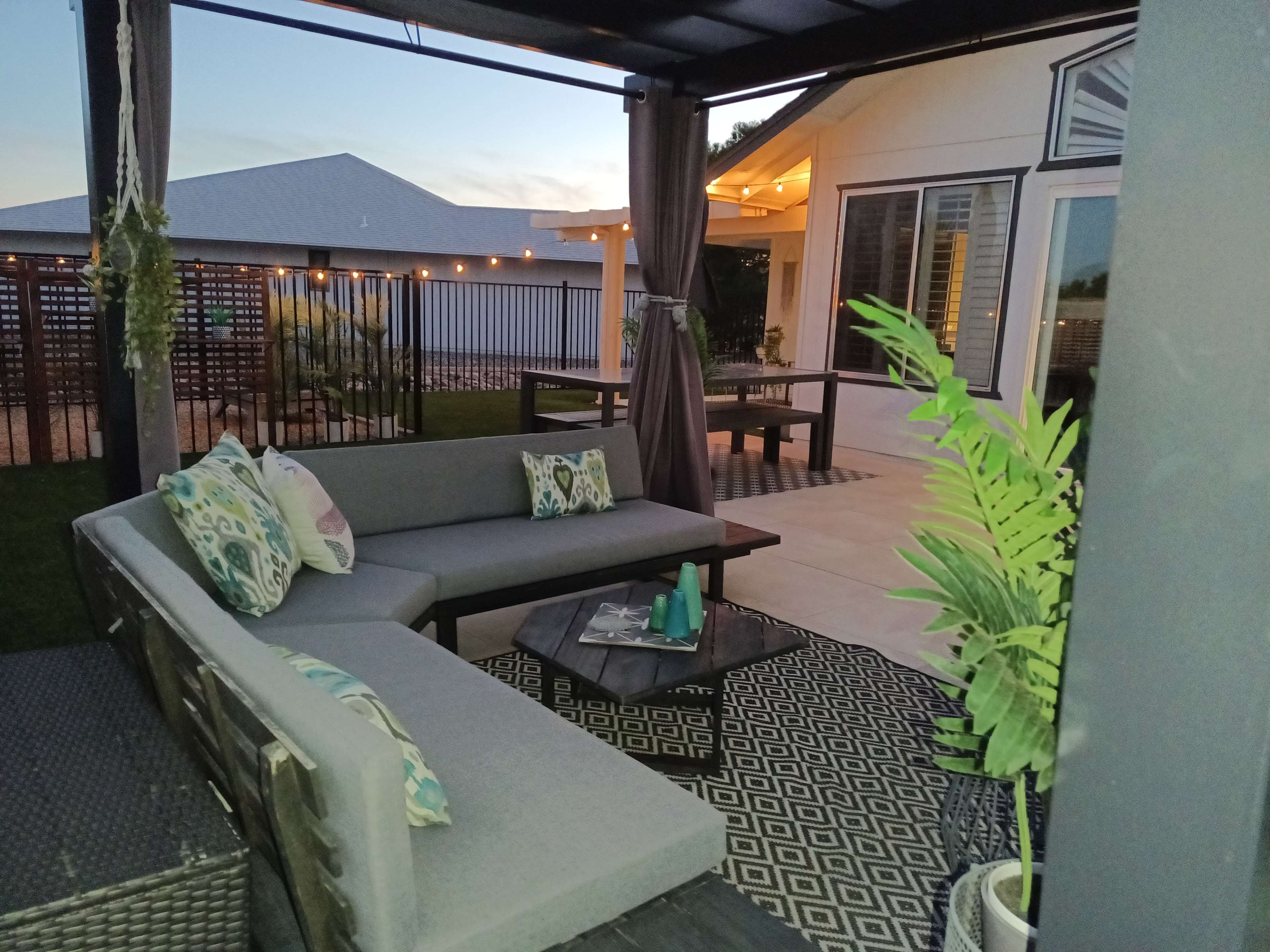 A modern outdoor seating area features a sectional sofa with decorative pillows, a small table, and plants, all under a covered pavilion.