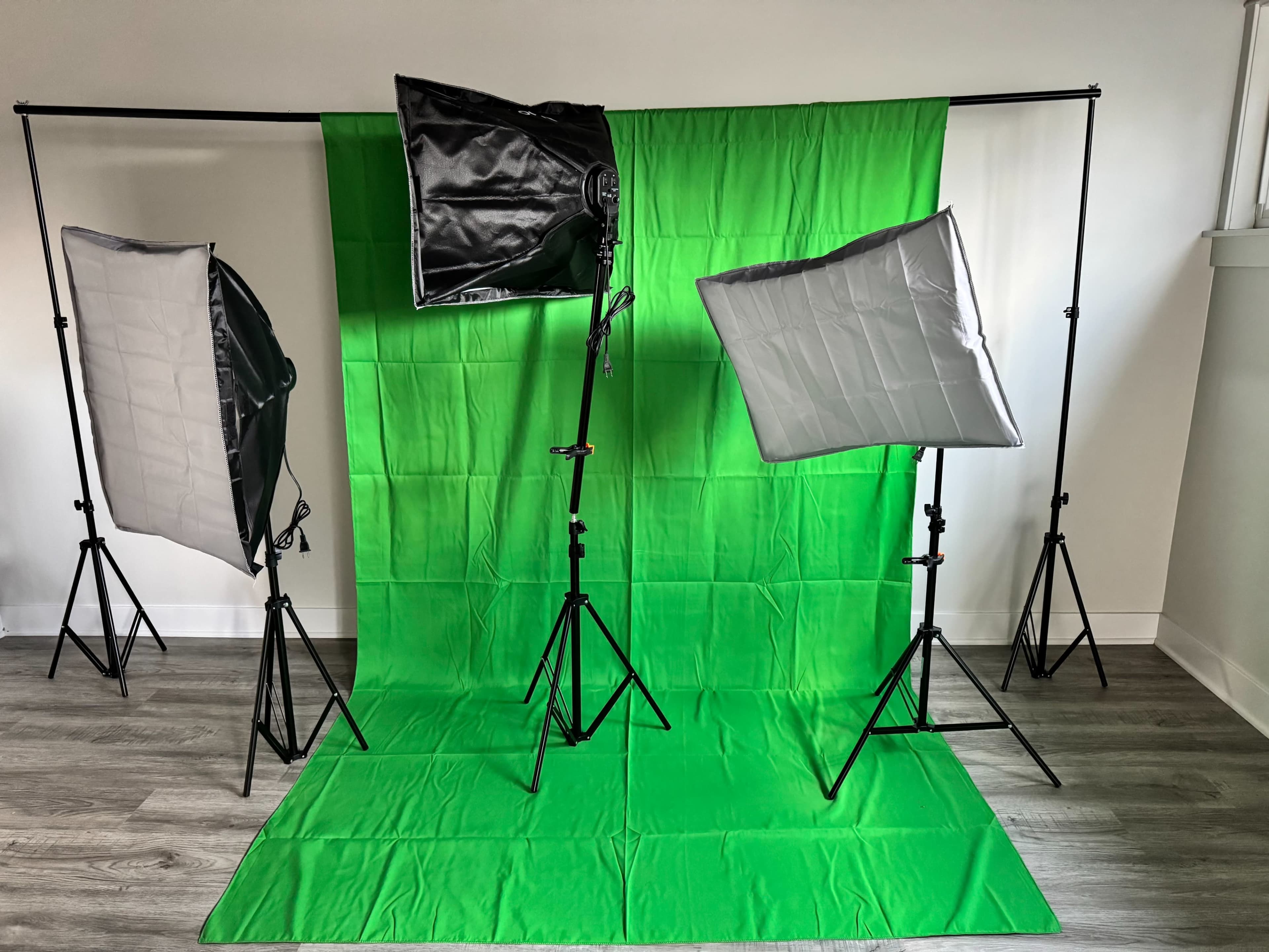 The image shows a photography setup featuring three softbox lights and a large green backdrop on a wooden floor.