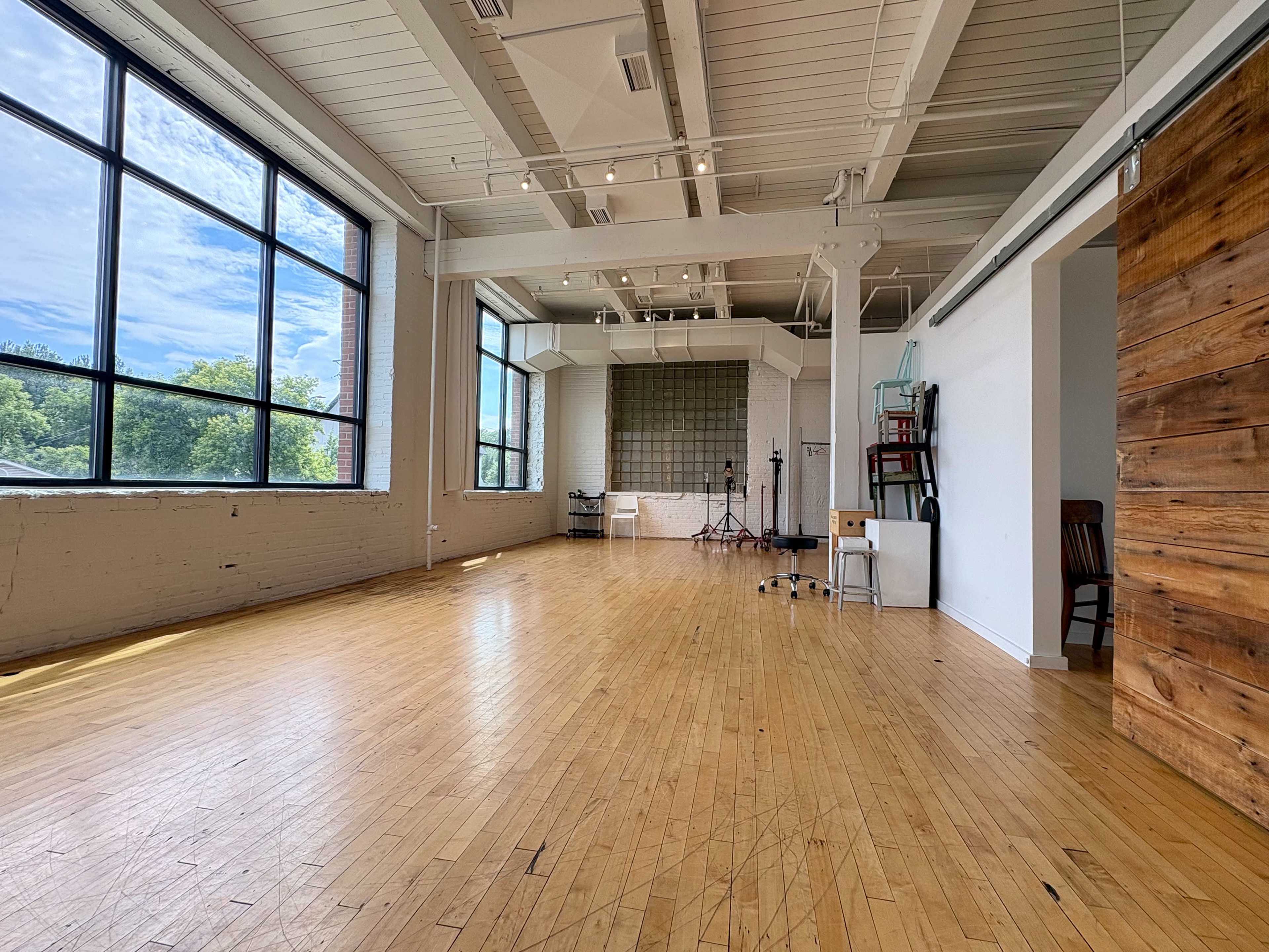 The image shows a spacious, well-lit interior with wooden floors, large windows, and a simple backdrop set up for photography or filming.