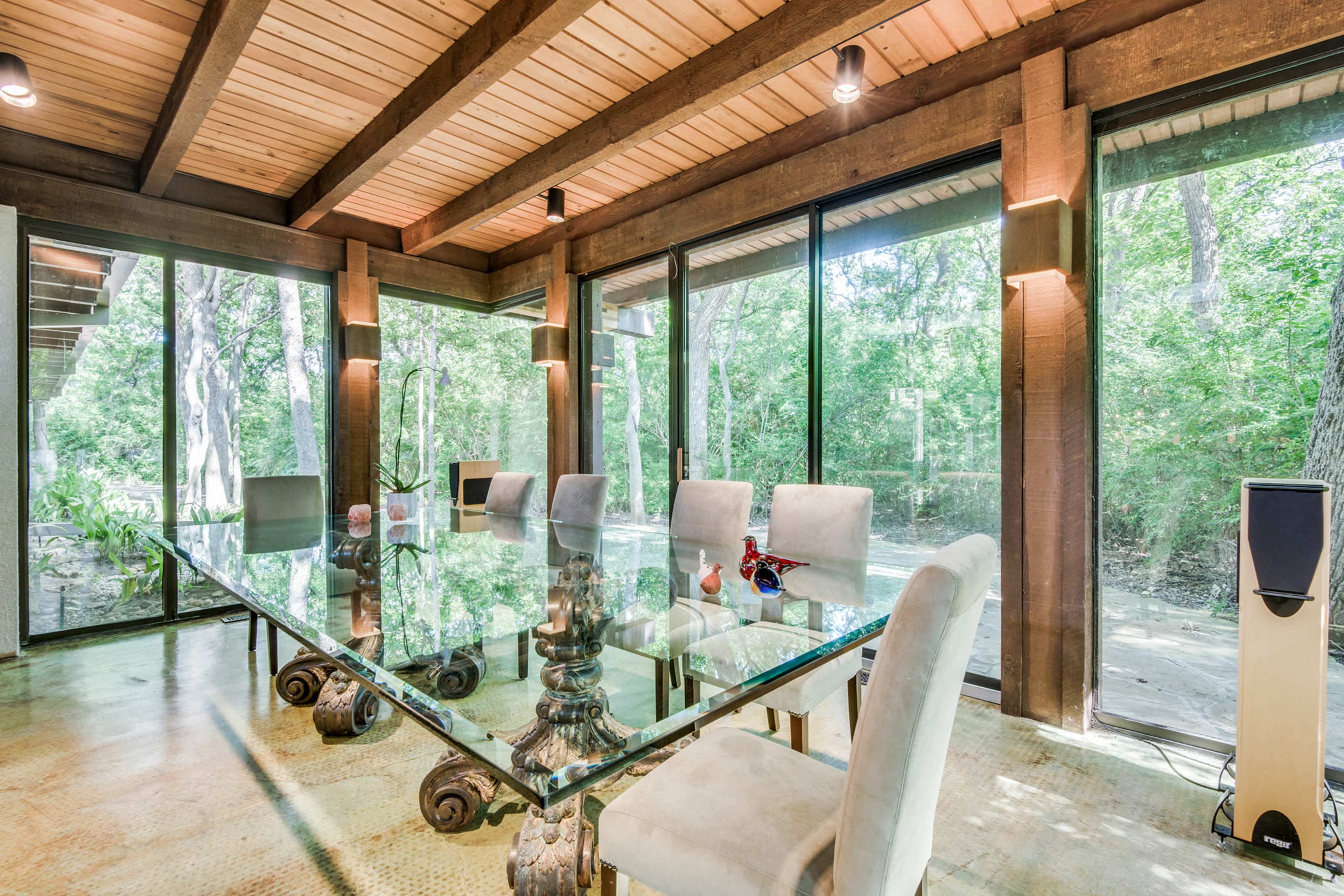 A modern conference room features a large glass table surrounded by upholstered chairs, with floor-to-ceiling windows offering views of a wooded landscape.