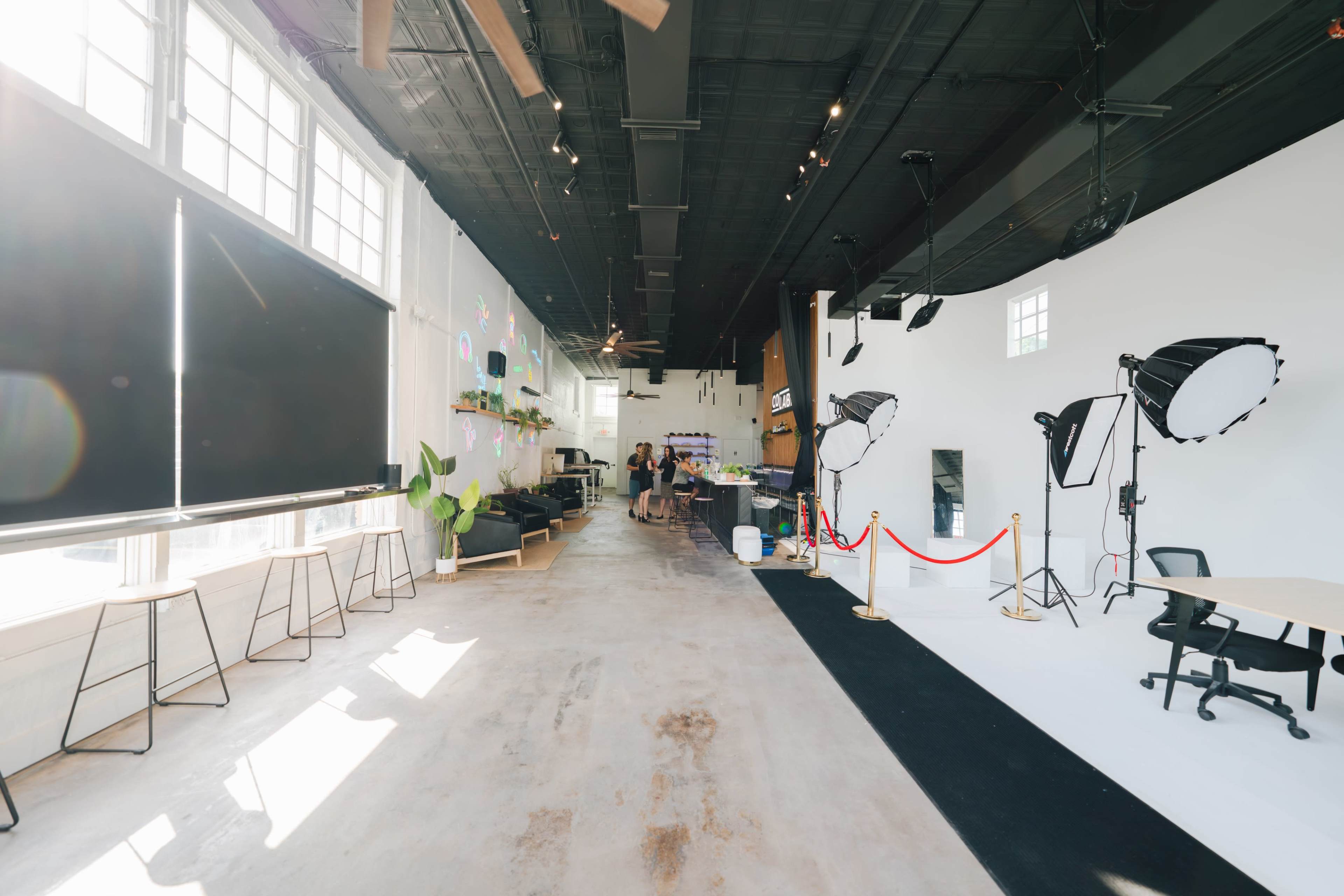 A spacious studio with large windows, plants, and multiple photography lighting setups, featuring a black carpet and a reception area.