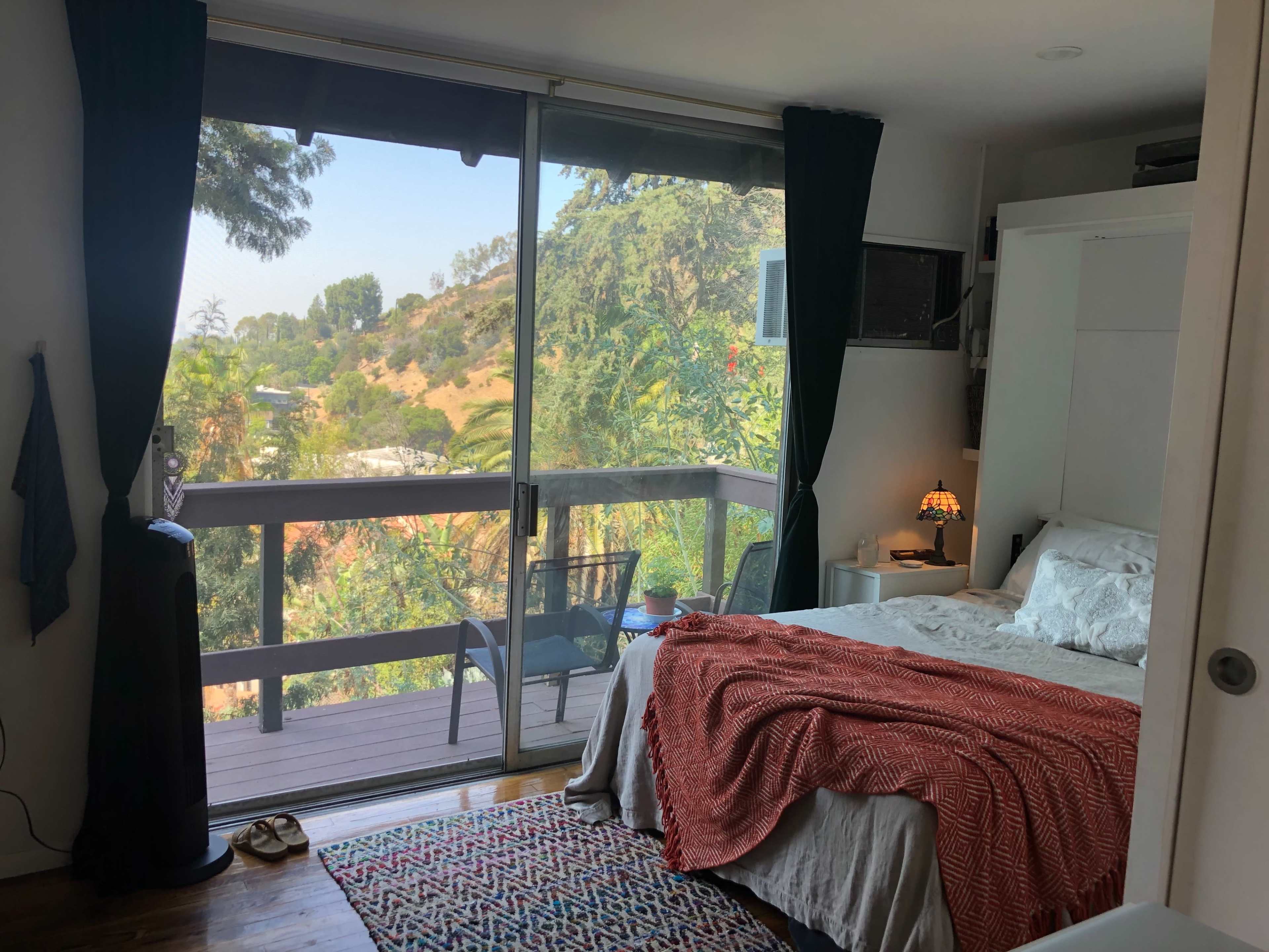 A bedroom features a bed with a patterned blanket, a TV mounted on the wall, and large sliding glass doors that open to a view of green hills.