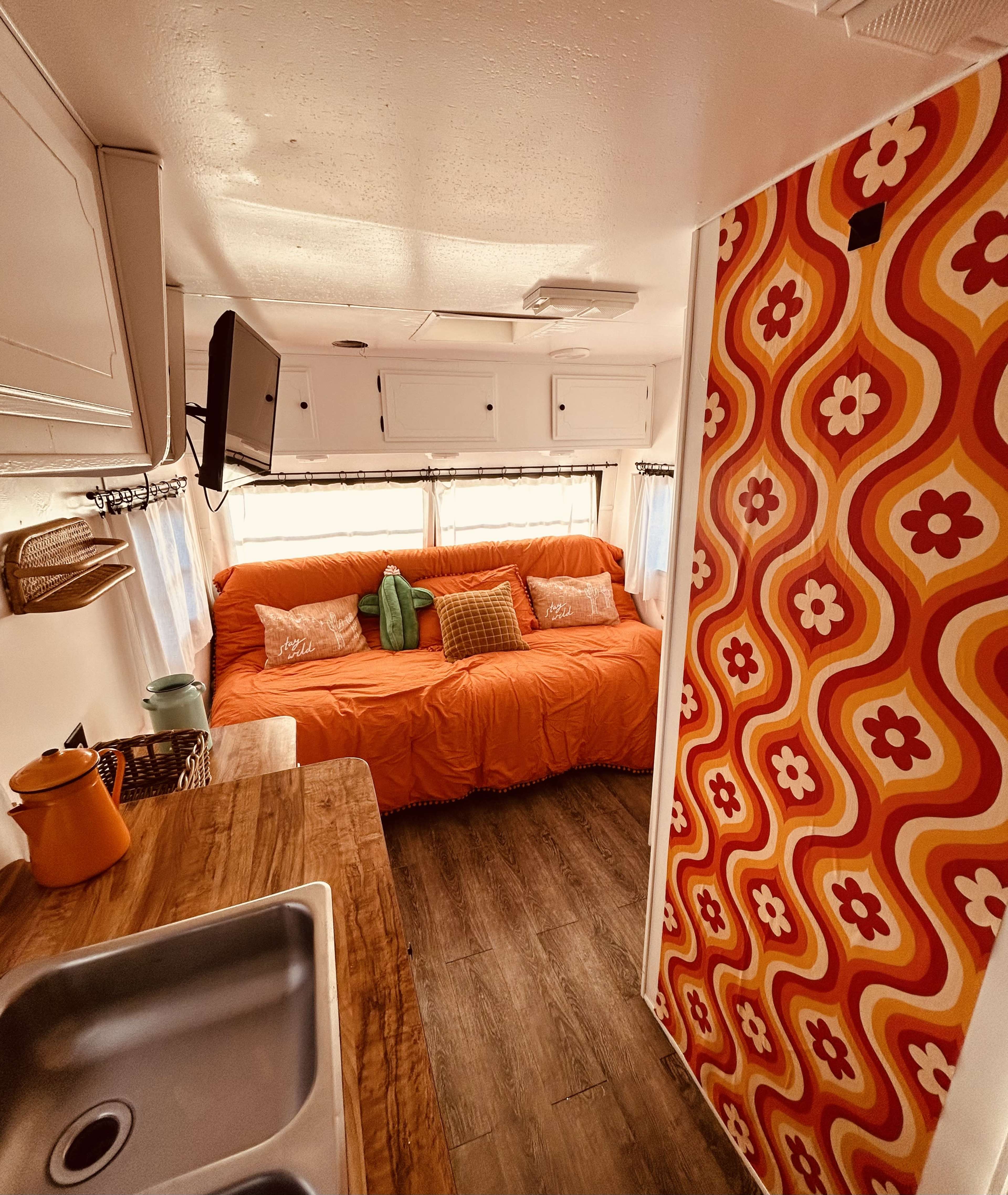 The interior of a camper van featuring a bright orange sofa, a wooden table, and retro floral wallpaper.