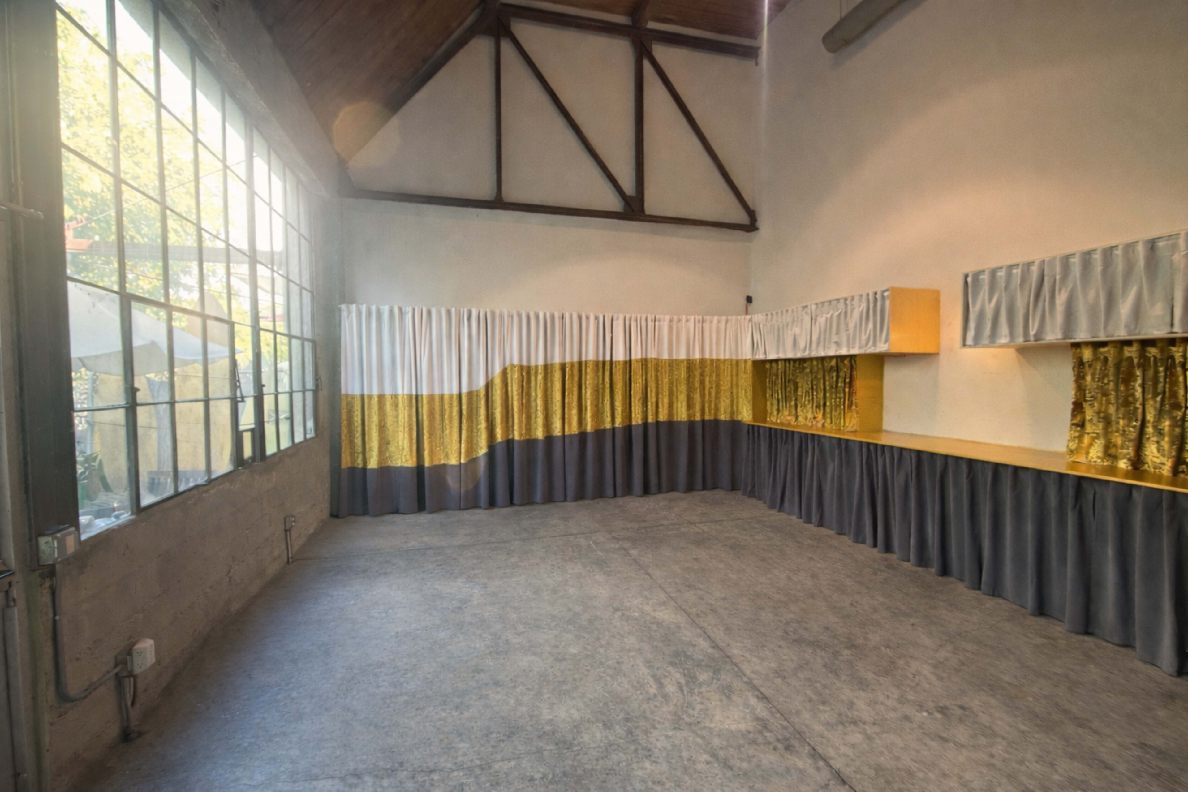 The image shows an empty room with large windows, featuring a textured floor and walls adorned with fabric curtains in gold and gray tones.