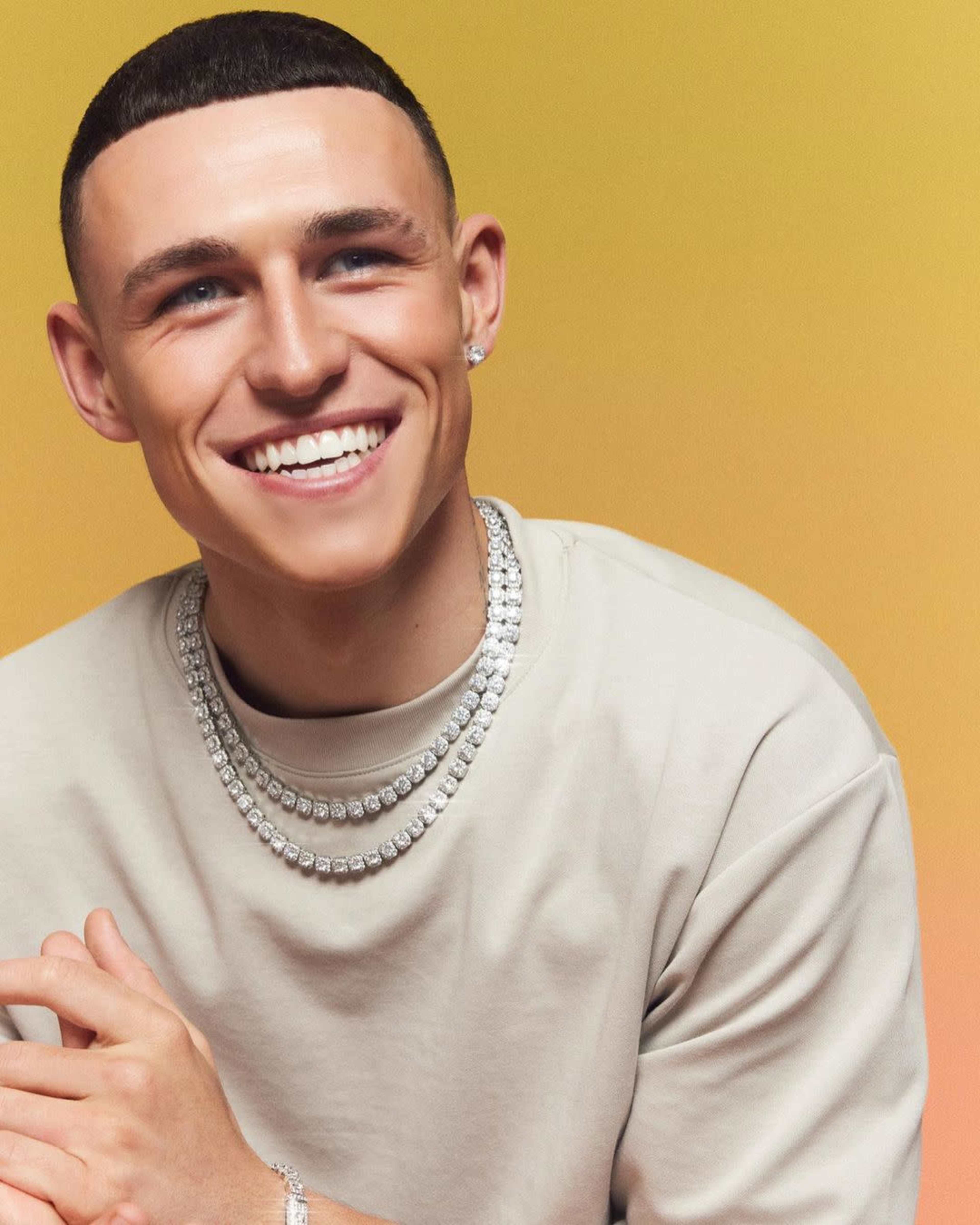 A man with a short hairstyle smiles while wearing a light-colored shirt and multiple beaded necklaces against a gradient yellow background.