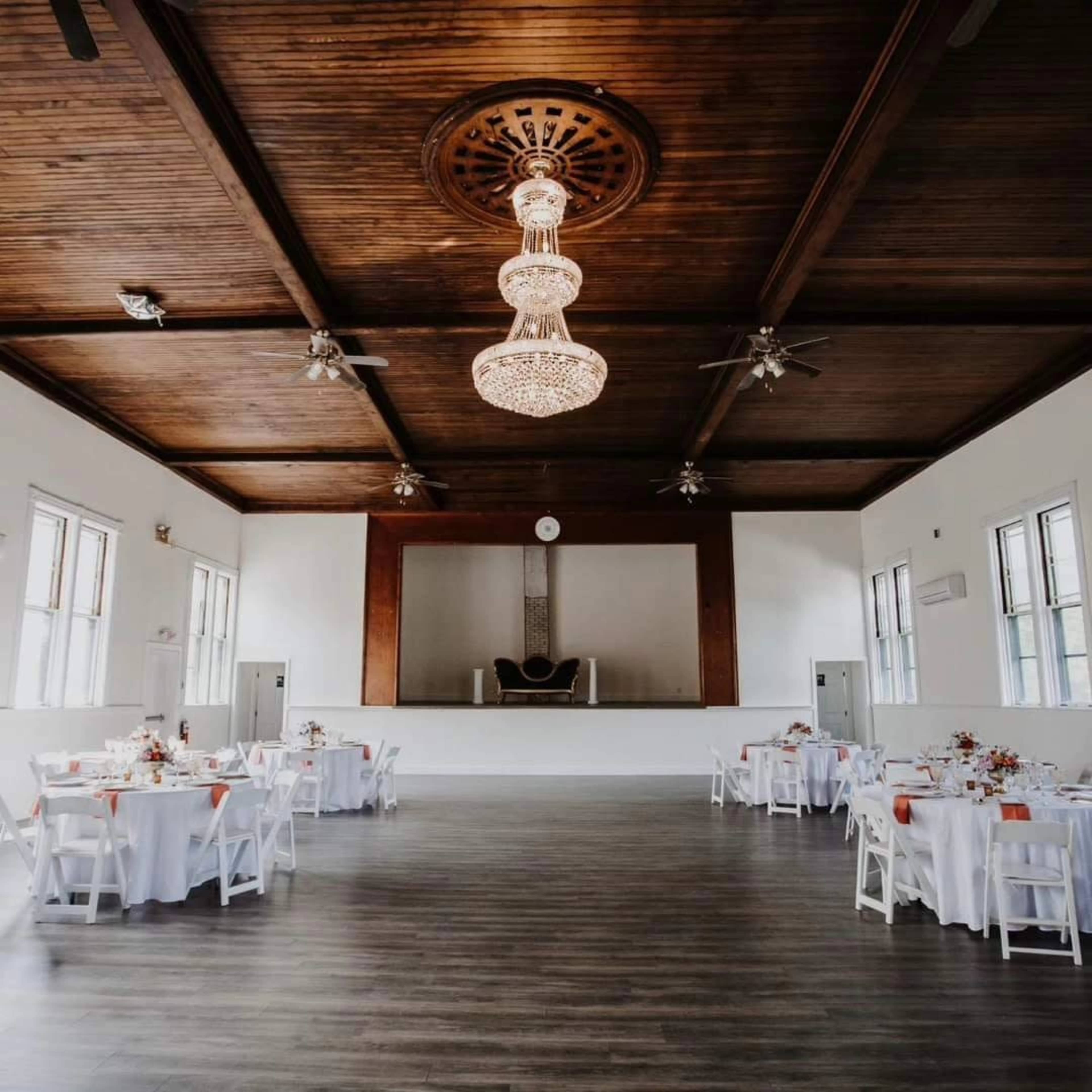 The image depicts a spacious event hall with white tables set for dining, a wooden ceiling adorned with a chandelier, and a stage at the far end.
