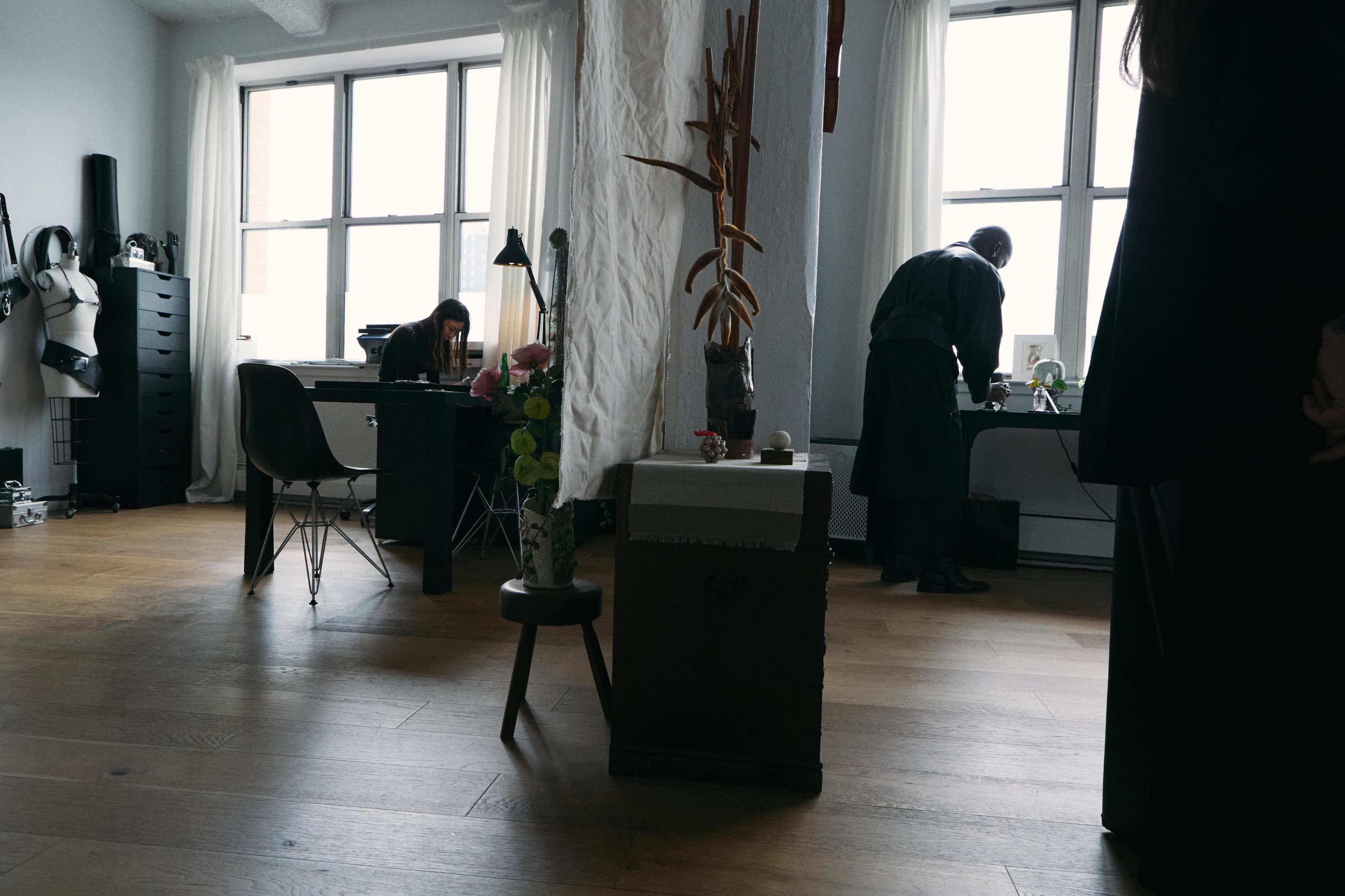 The image shows a modern office space with two people working at desks, large windows letting in natural light, and a decorative plant in the foreground.