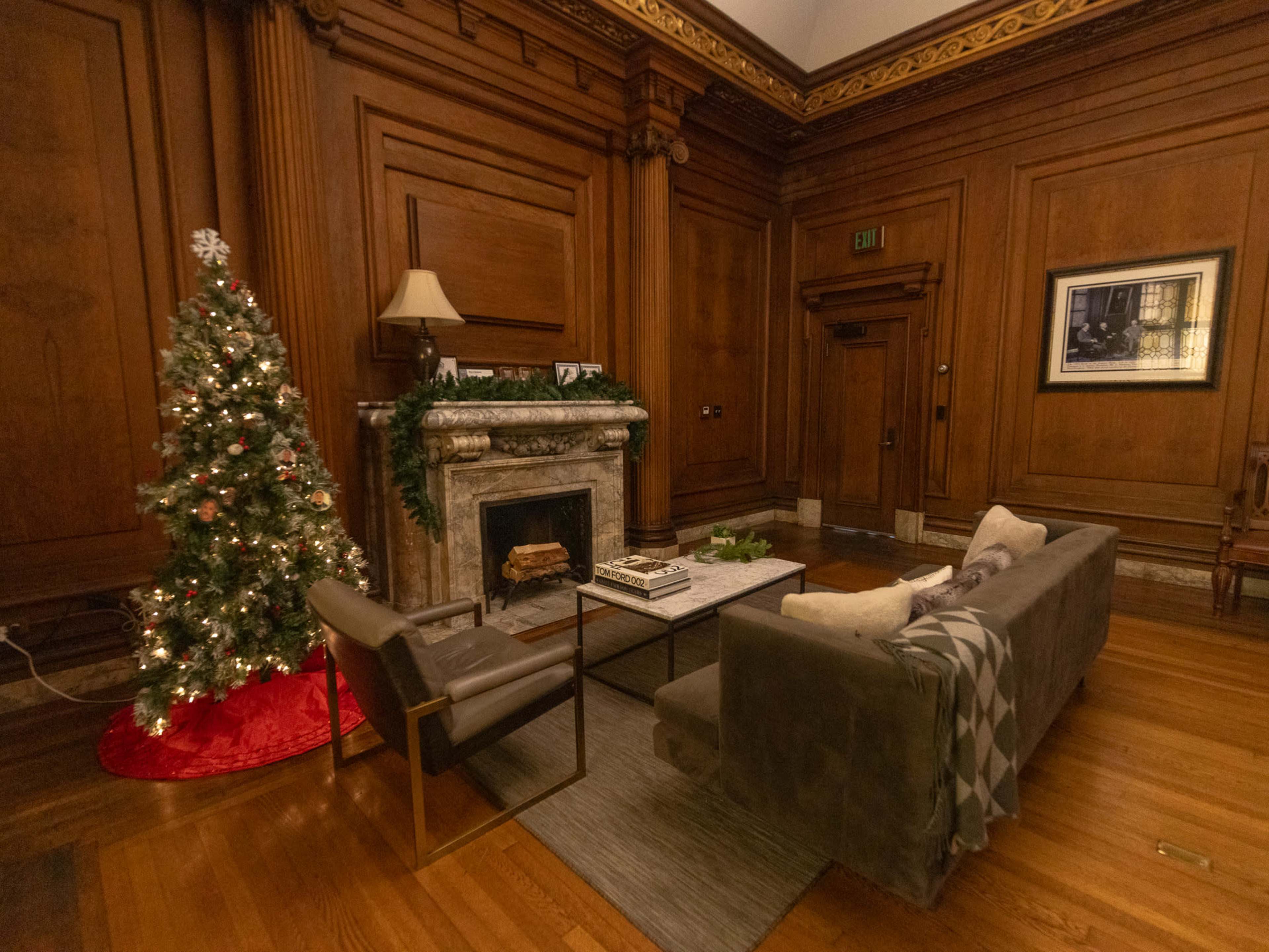 A decorated Christmas tree stands next to a fireplace in a cozy living room with rich wooden paneling and elegant furnishings.