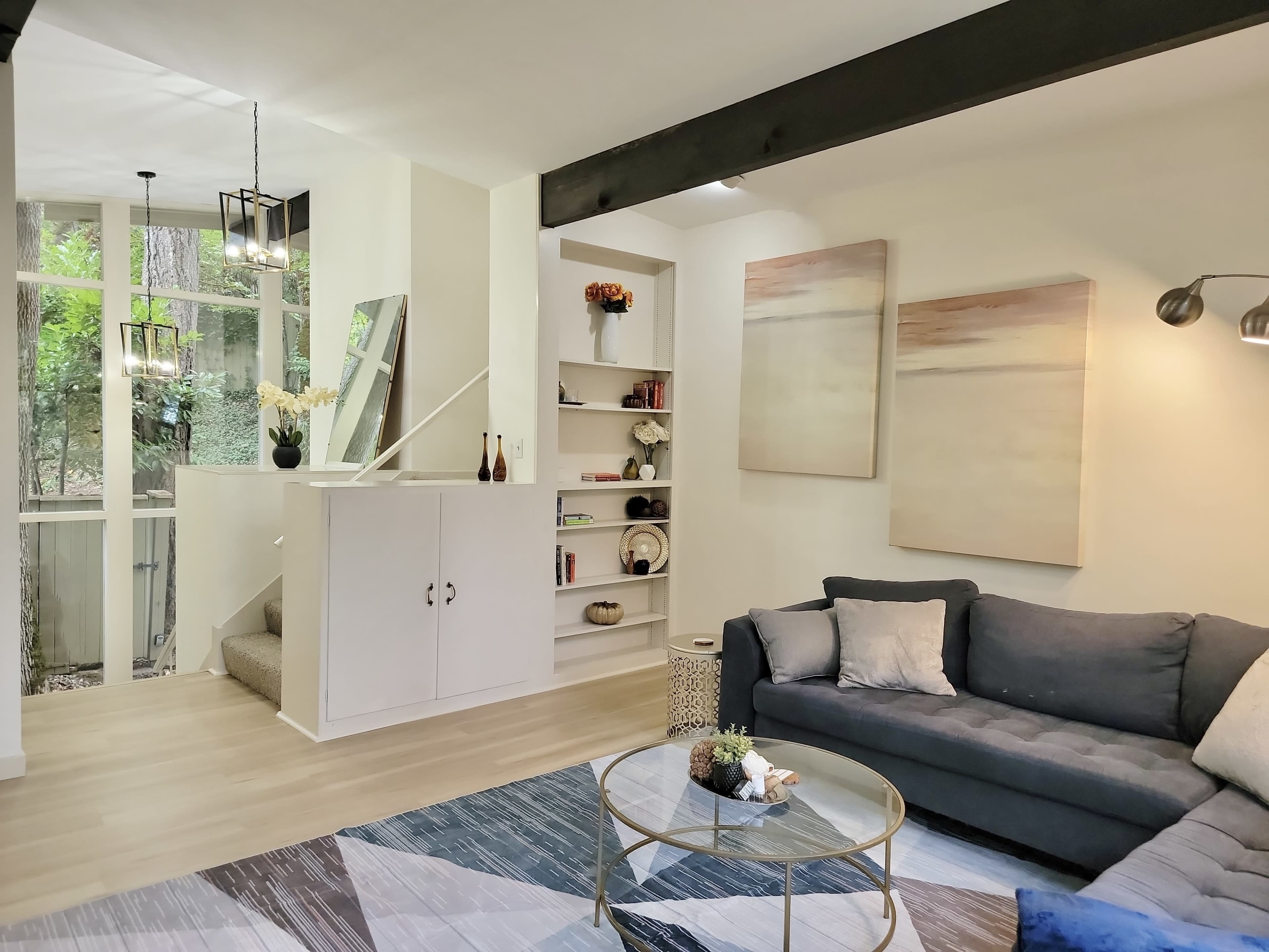 The image shows a modern living room with a blue sofa, a round coffee table, and a staircase leading to an upper level, surrounded by light-colored walls and natural light from large windows.