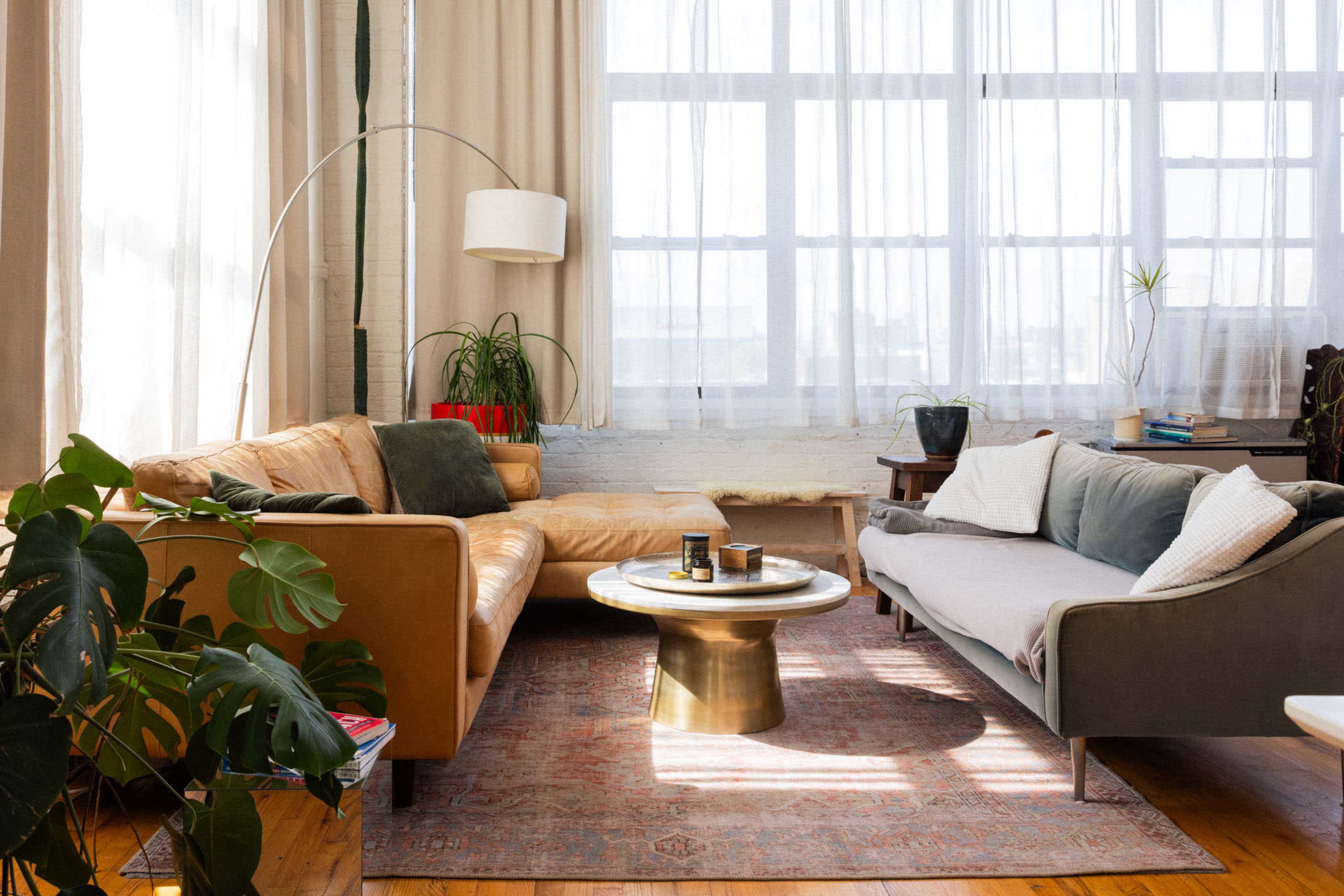The image features a bright living room with large windows, a beige sofa, a gray couch, and a round coffee table on a patterned rug.