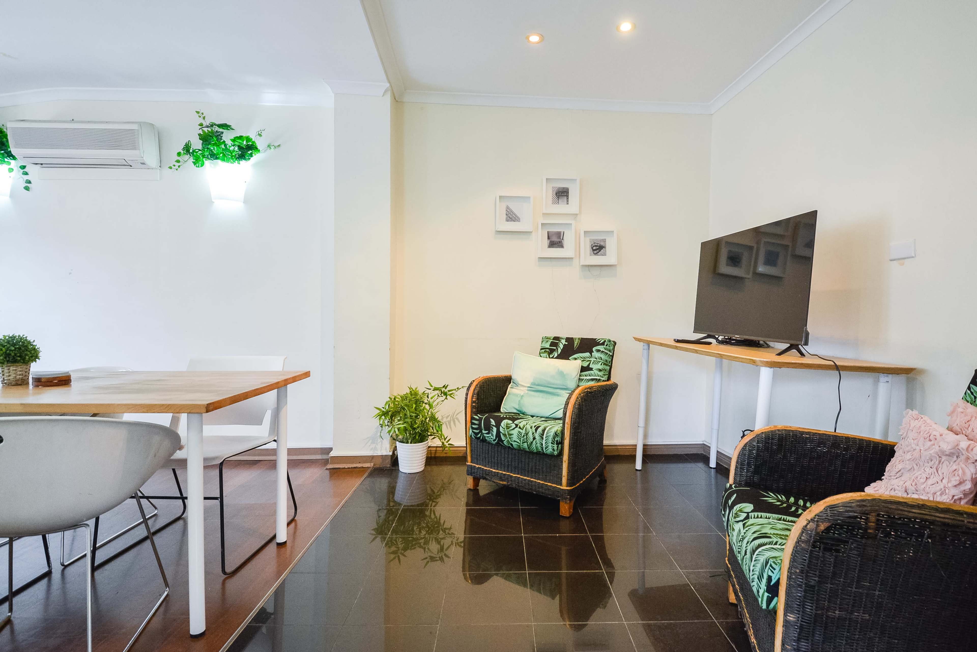 A cozy living area with a wooden table, two chairs, and a television mounted on the wall, complemented by potted plants and framed pictures.