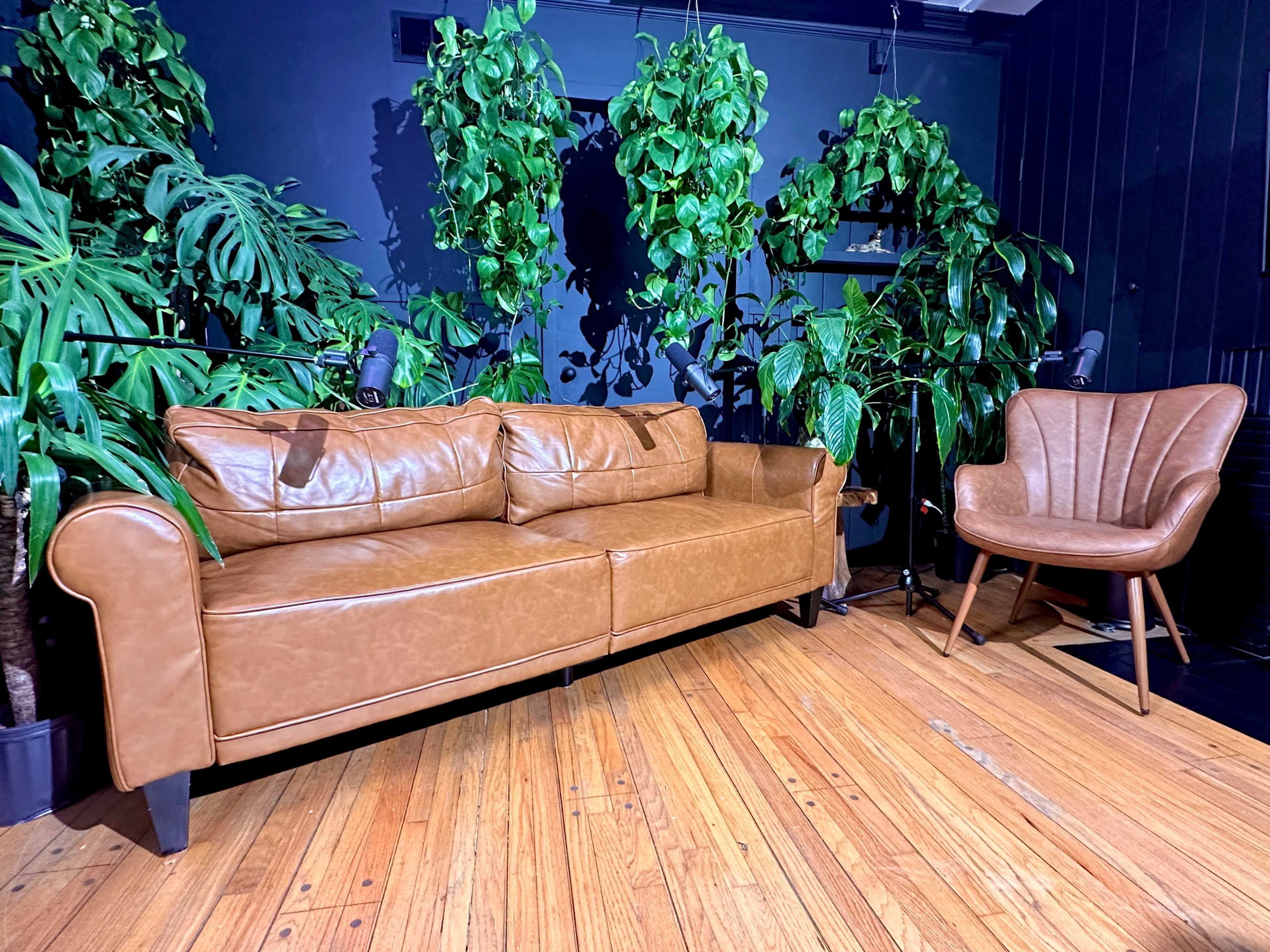 A brown leather couch is positioned next to a matching chair in a room filled with various green plants against a dark wall.